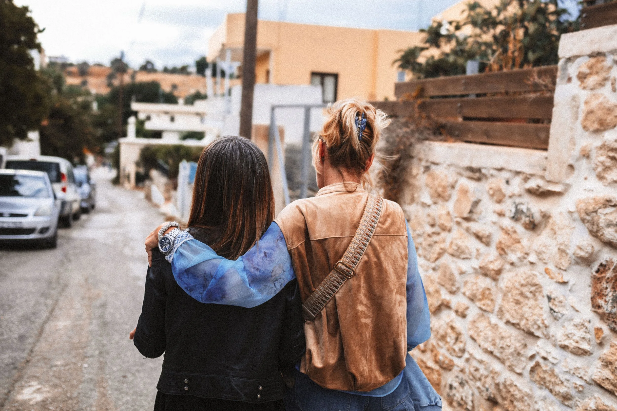 Zwei Frauen gehen Arm in Arm durch eine Straße in einer Wohngegend, eine trägt einen braunen Rucksack, die andere hat lange dunkle Haare.