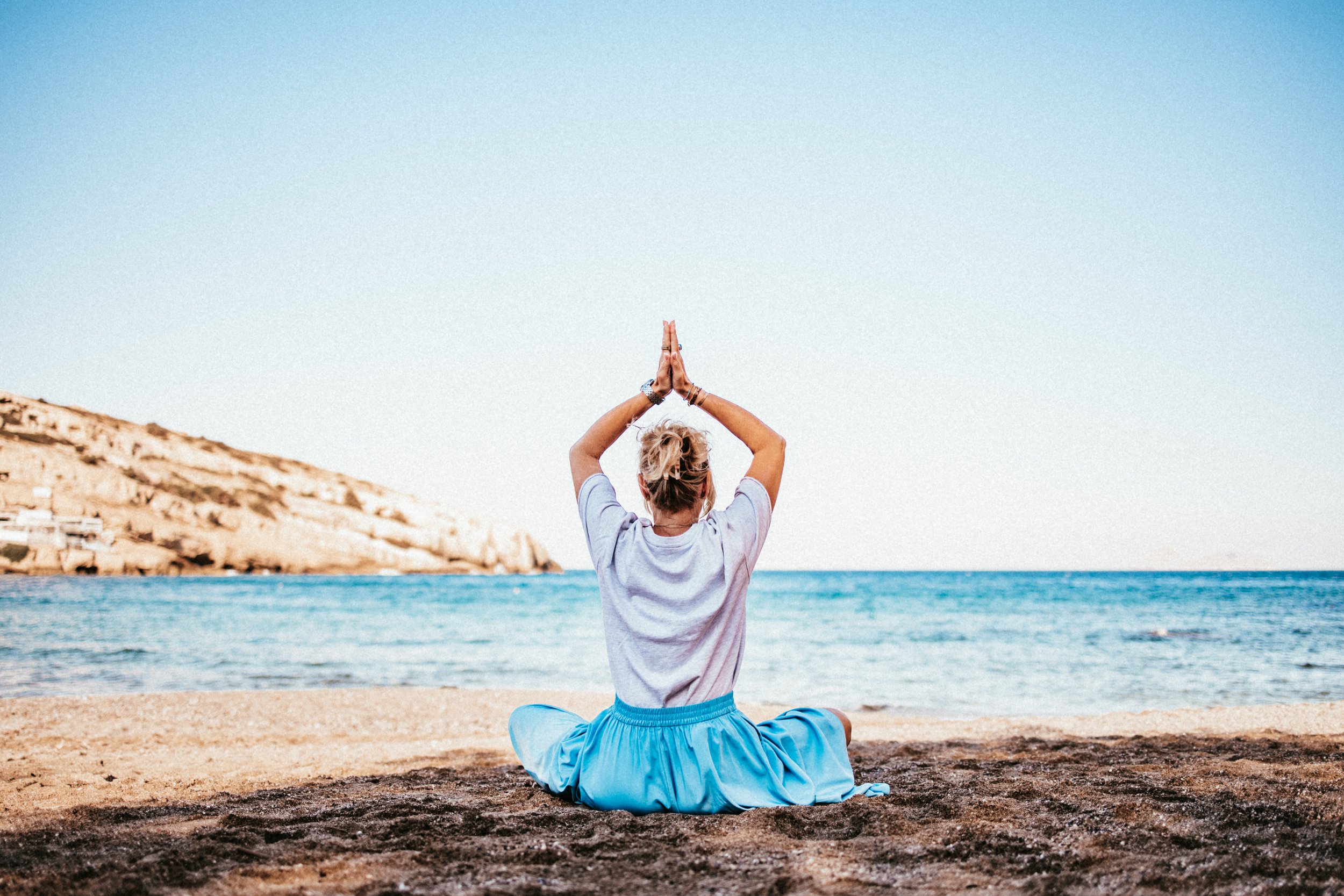 Eine Person sitzt im Schneidersitz am Strand, macht Yoga und blickt auf das Meer.
