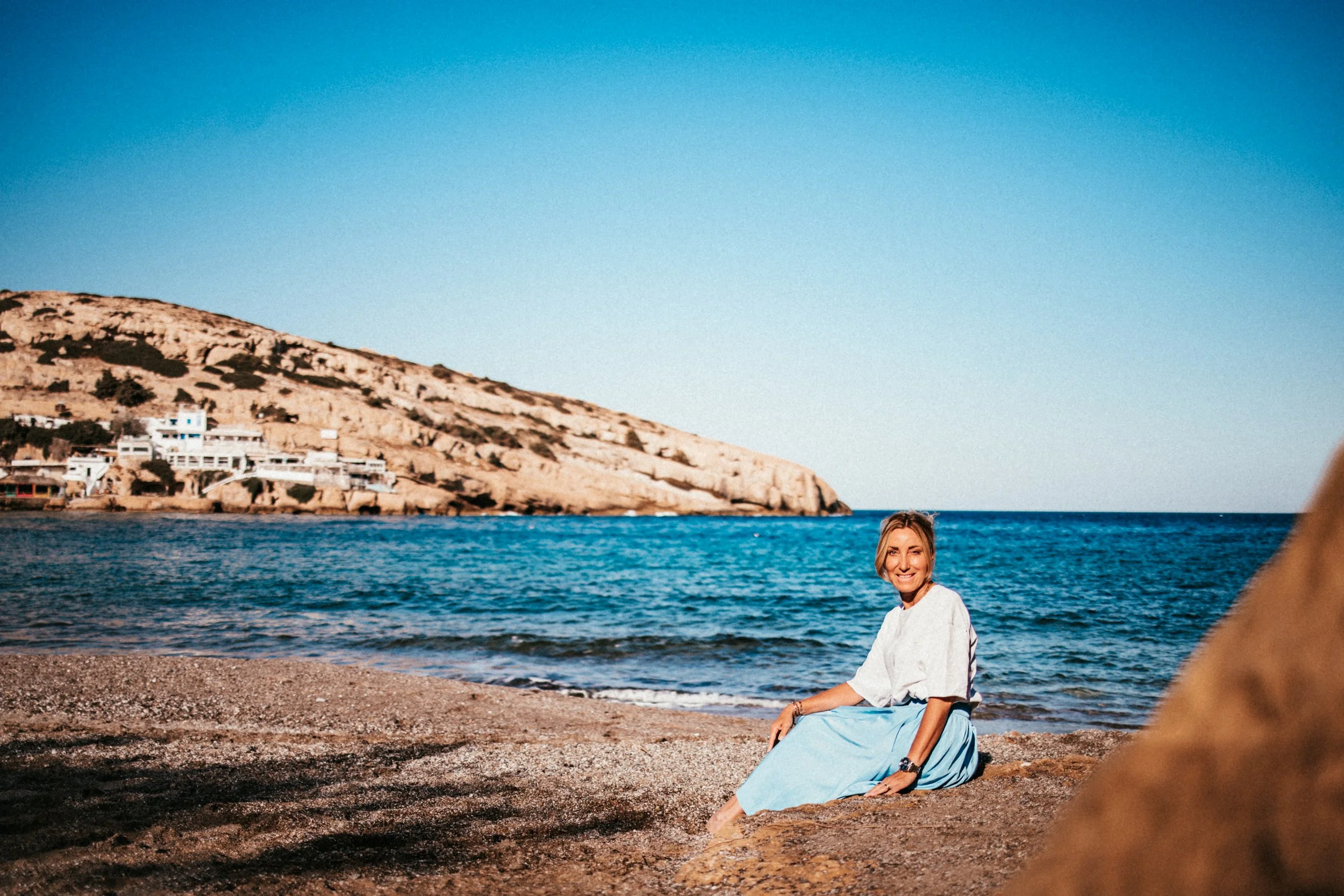 Eine Frau sitzt entspannt am Strand, lächelnd, vor dem blauen Meer und einem felsigen Hügel im Hintergrund.