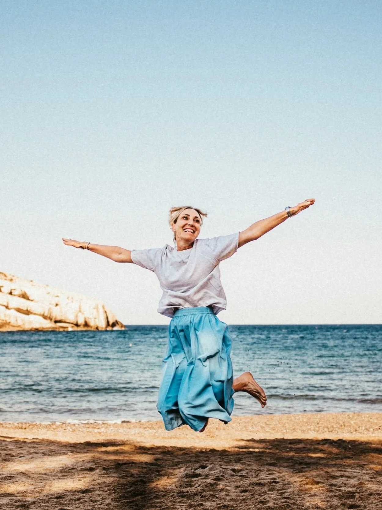 Eine lachende Frau springt an einem Strand. Sie trägt eine graue Bluse und eine blaue, weite Hose. Im Hintergrund ist das Meer und ein Felsen zu sehen.
