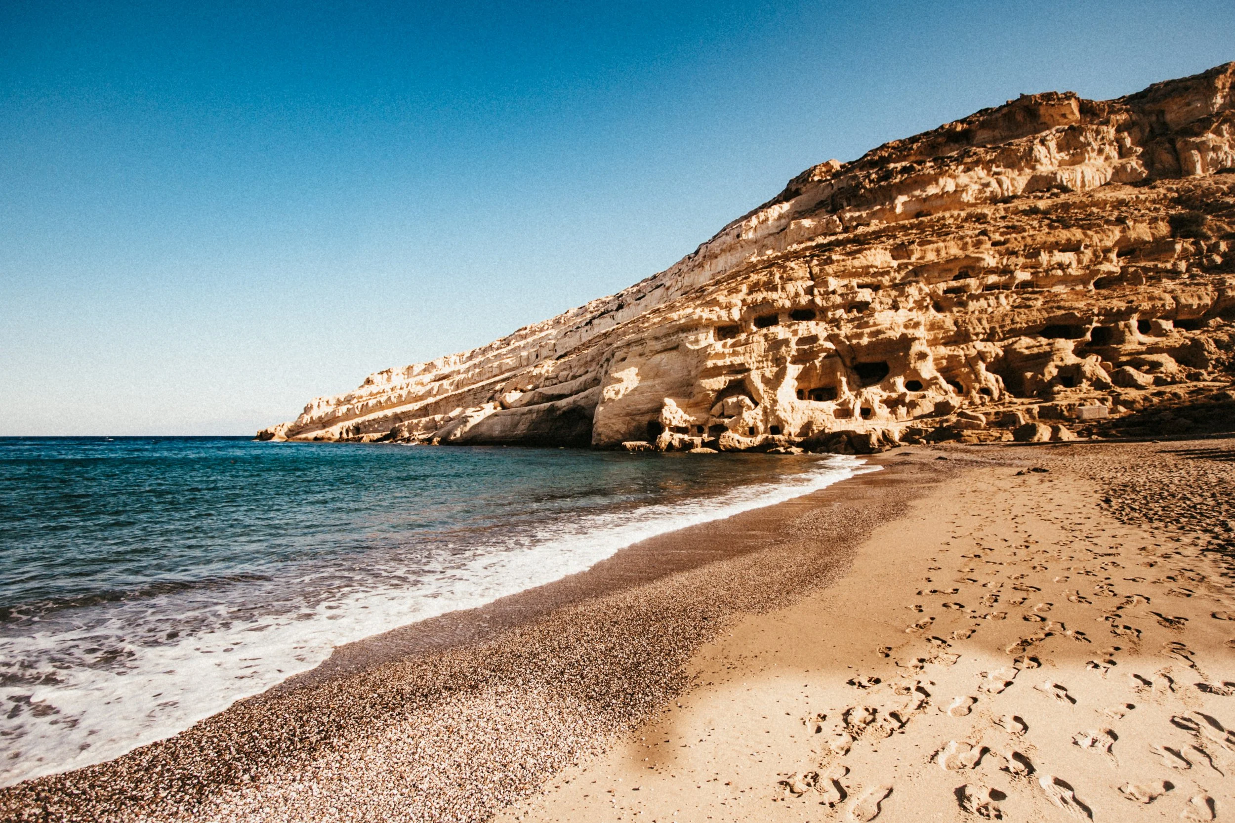 Strand mit sandigem Ufer, Klippen mit Höhlen im Hintergrund und blauer Himmel.
