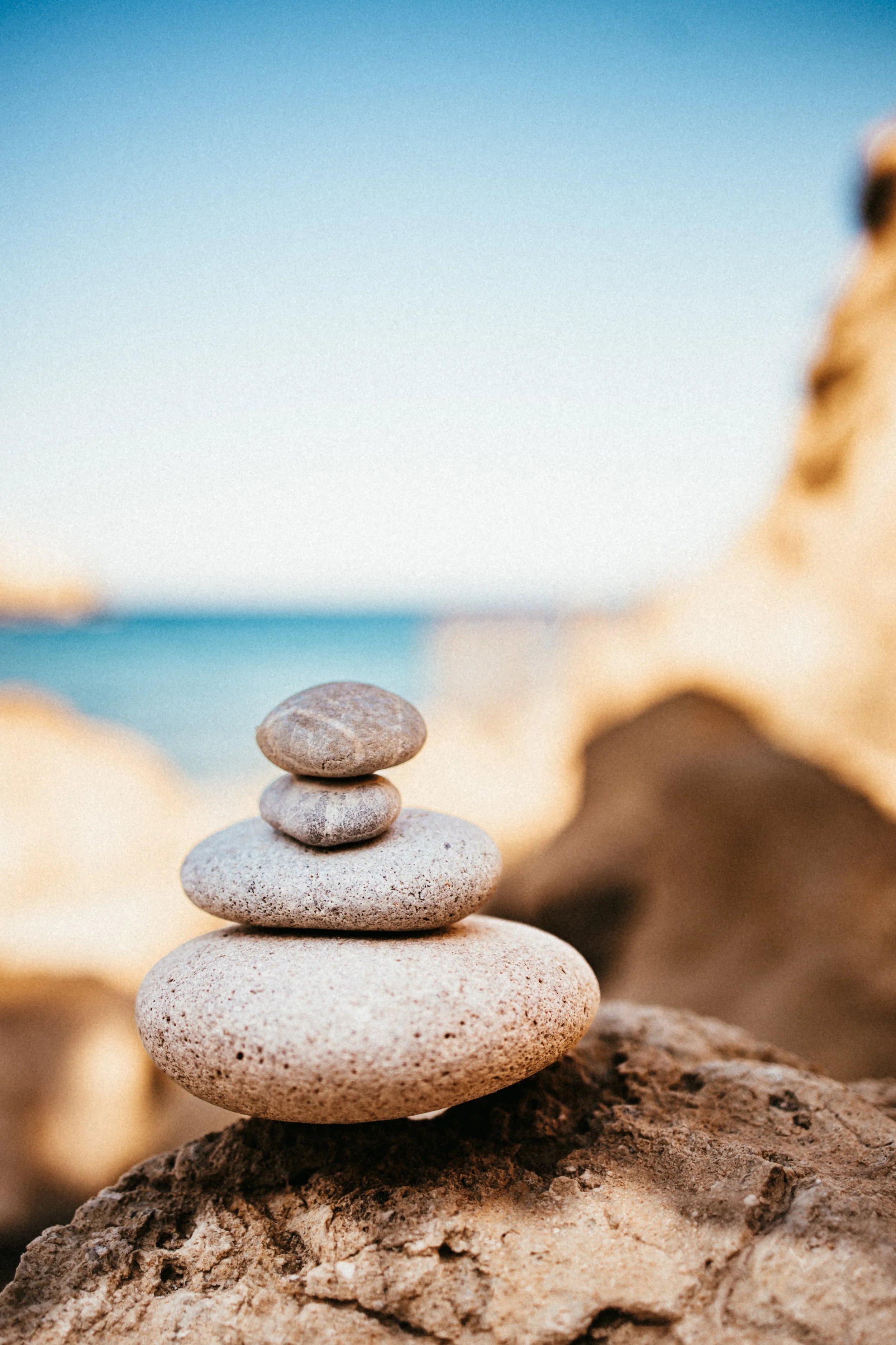 Fünf flache Steine, die auf einem größeren Stein gestapelt sind, am Strand bei Sonnenuntergang mit Blick auf das Meer im Hintergrund.