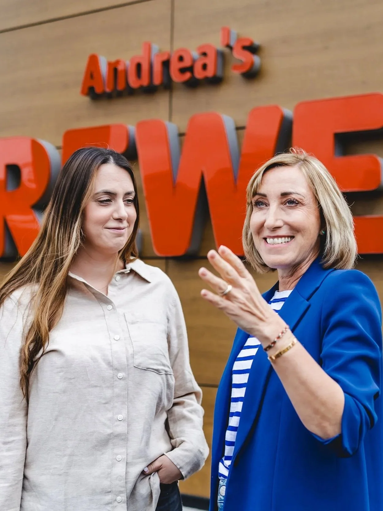 Zwei Frauen im Gespräch, eine lächelnd, die andere wirkt nachdenklich, vor einem Holz-Wand mit großen roten Buchstaben, die „Andrea’s REWE“ schreiben.