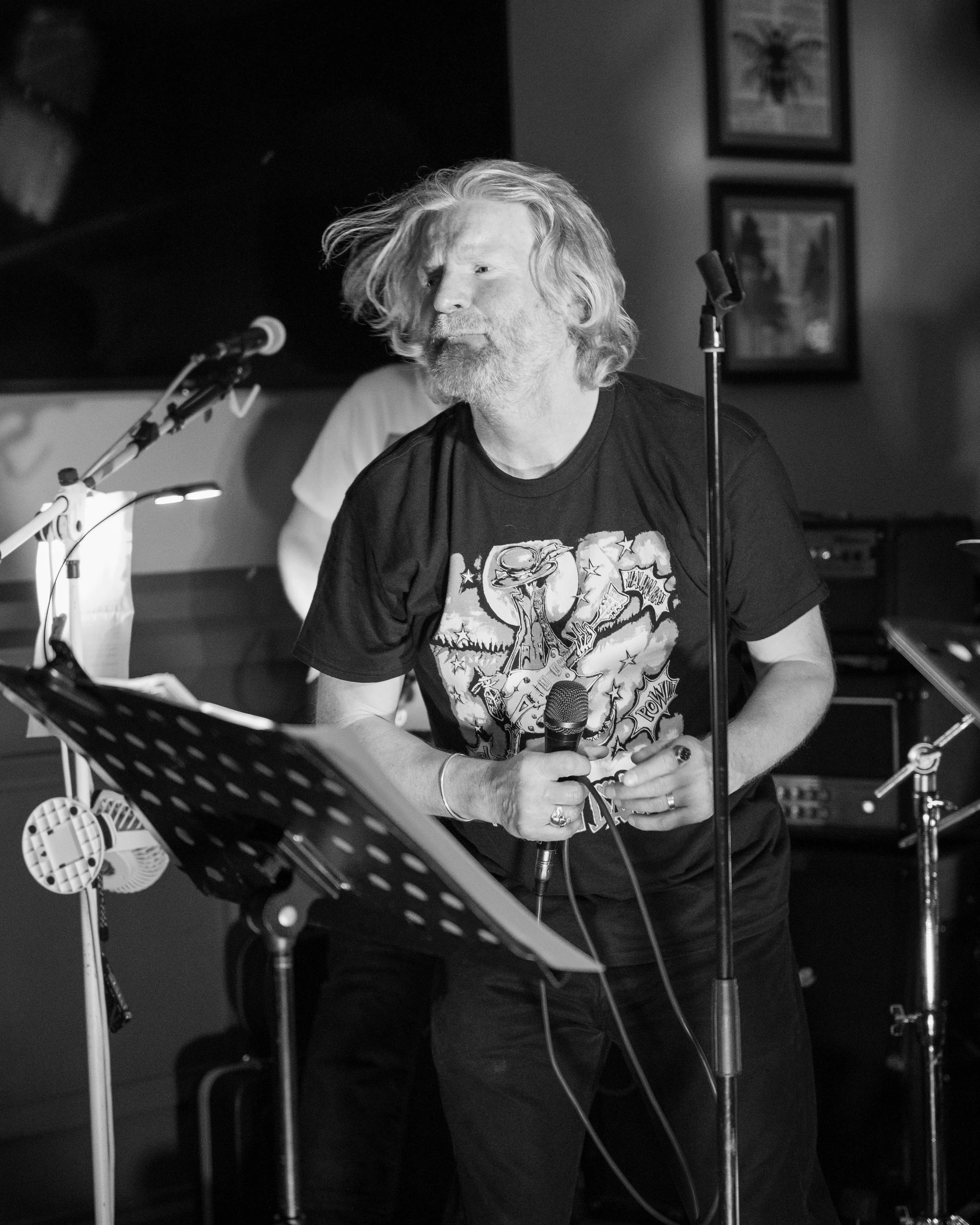 Man with long gray hair and beard singing into a microphone, wearing a graphic T-shirt, in a dimly lit bar or music venue.