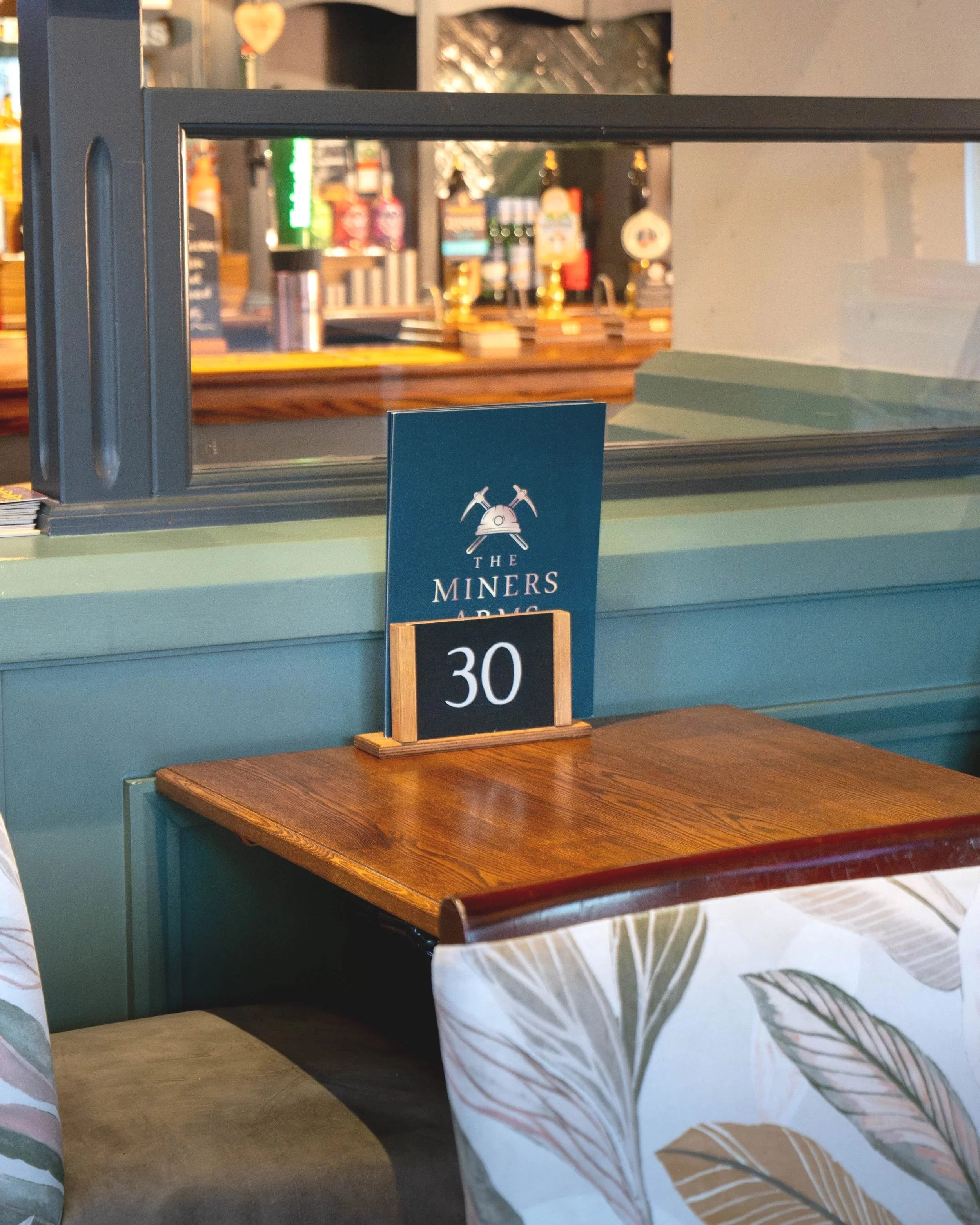 A wooden table with a blackboard displaying the number 30, situated in front of a teal-colored wall with a sign for The Miners Arms. Chairs with patterned cushions are partially visible in the foreground, and the background shows a bar area with bottles and taps.
