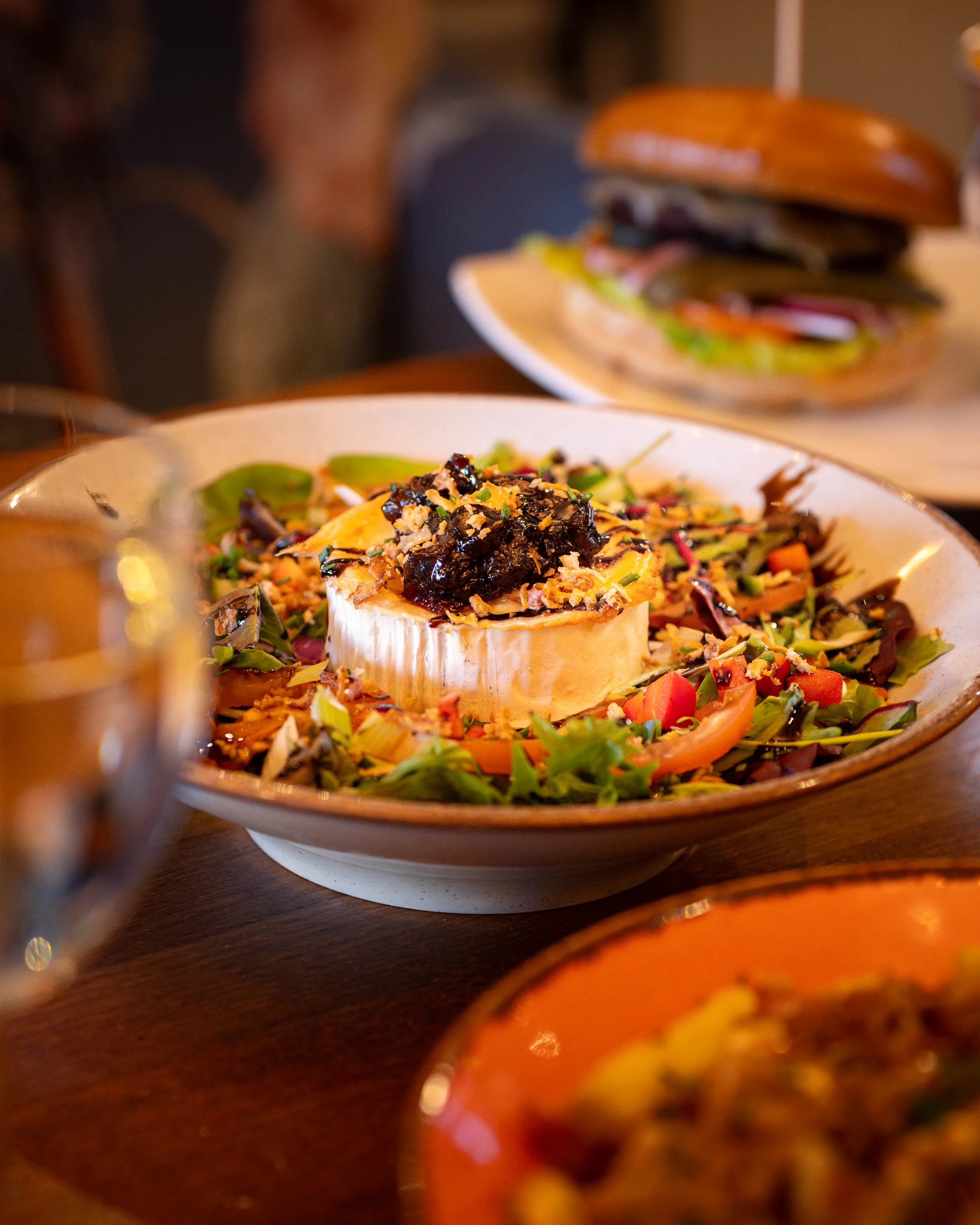 A salad with a round, soft cheese topped with dark sauce and garnished with chopped nuts, served on a bed of mixed greens and vegetables in a white bowl at a restaurant table.