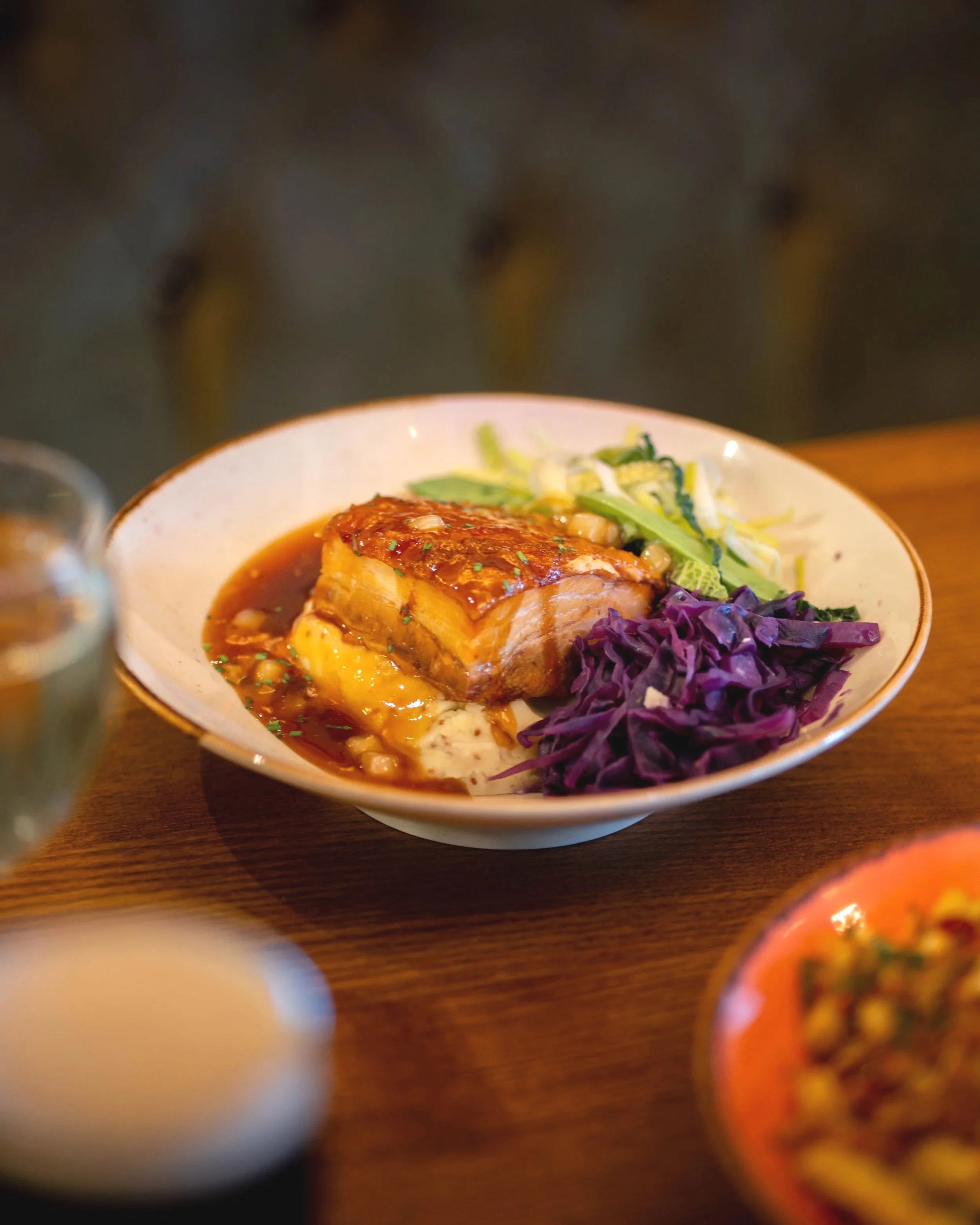 A bowl of meat with brown sauce, mashed potatoes, and purple cabbage, served with vegetables on the side.