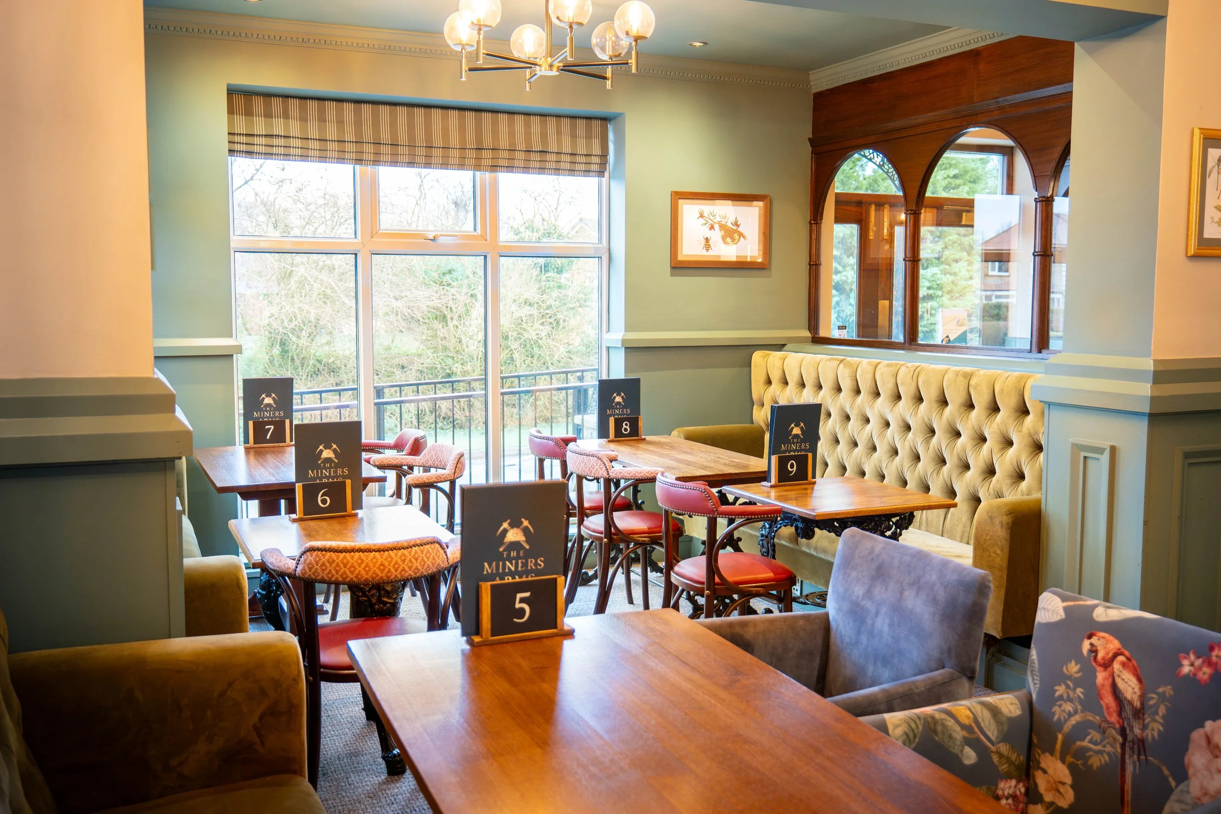 Interior of a cozy restaurant or café with various chairs and tables, some with numbered signs, large windows showing outdoor scenery, and decorative art on the walls.