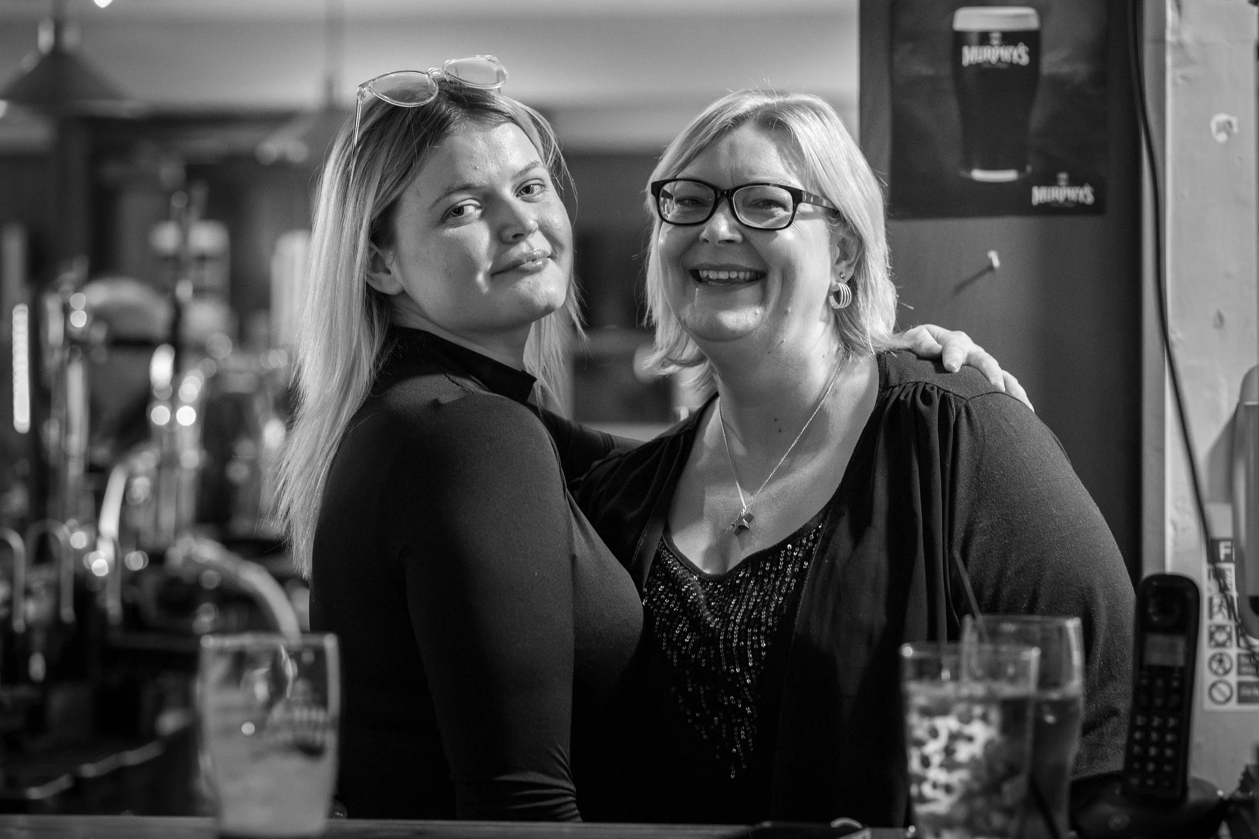 Two women smiling and posing together in a bar or restaurant, with drinks and bar equipment visible in the background.