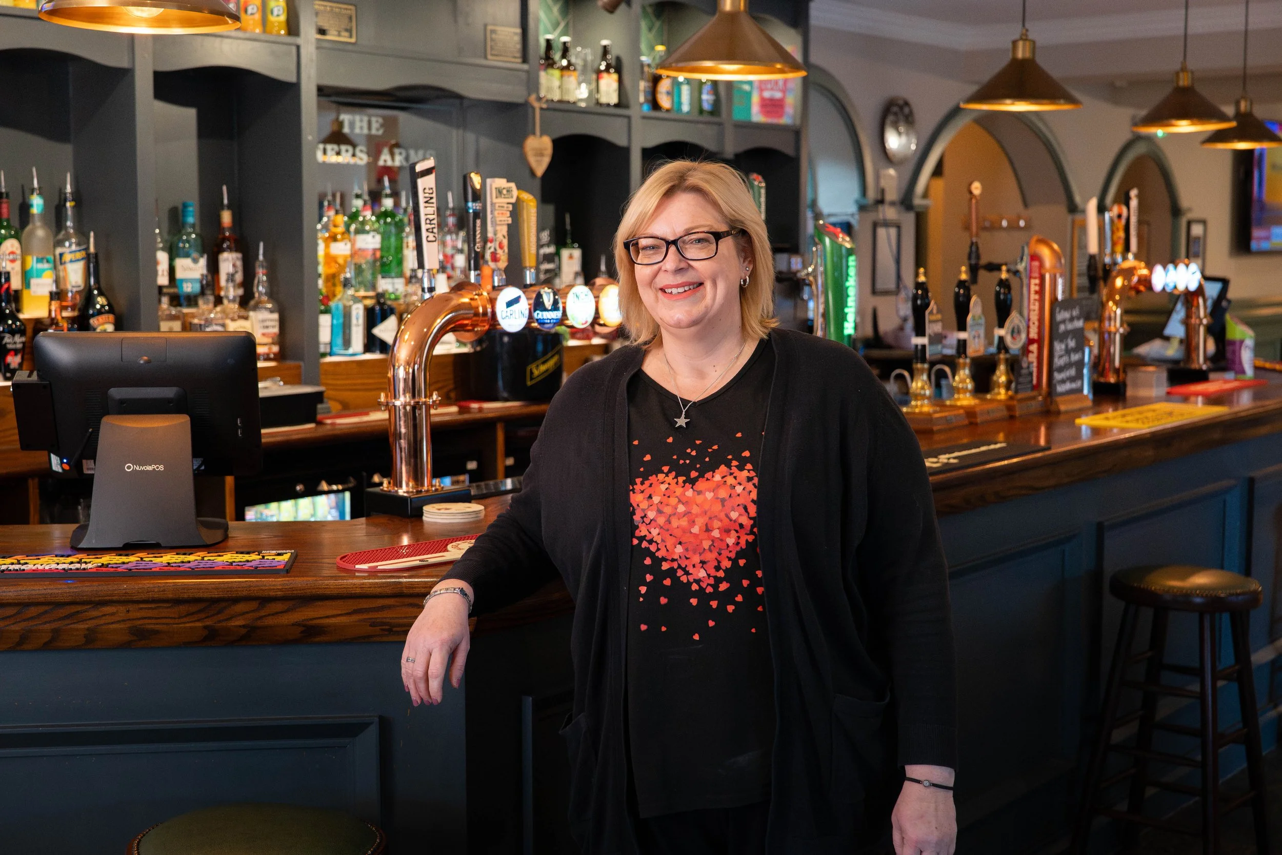 A woman with glasses and blonde hair in a black cardigan, standing at a bar with a smile, in a dimly lit pub with beer taps and bottles behind her.