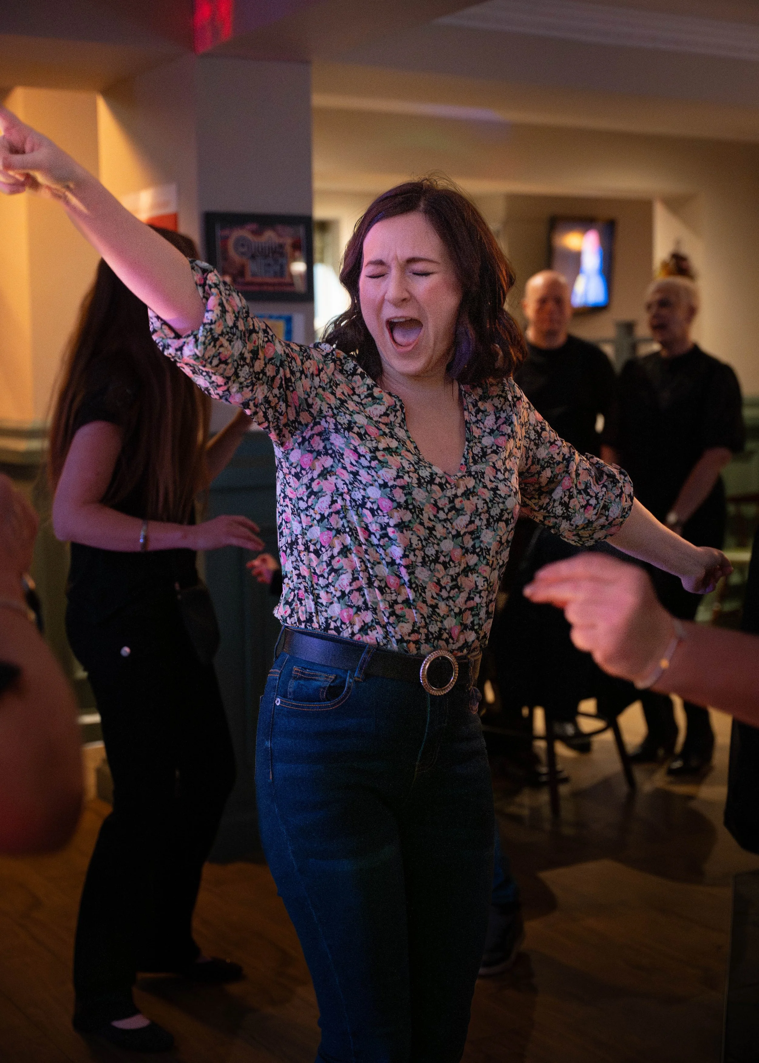A woman dancing and singing with eyes closed, wearing a floral blouse and dark jeans, in a dimly lit social setting.