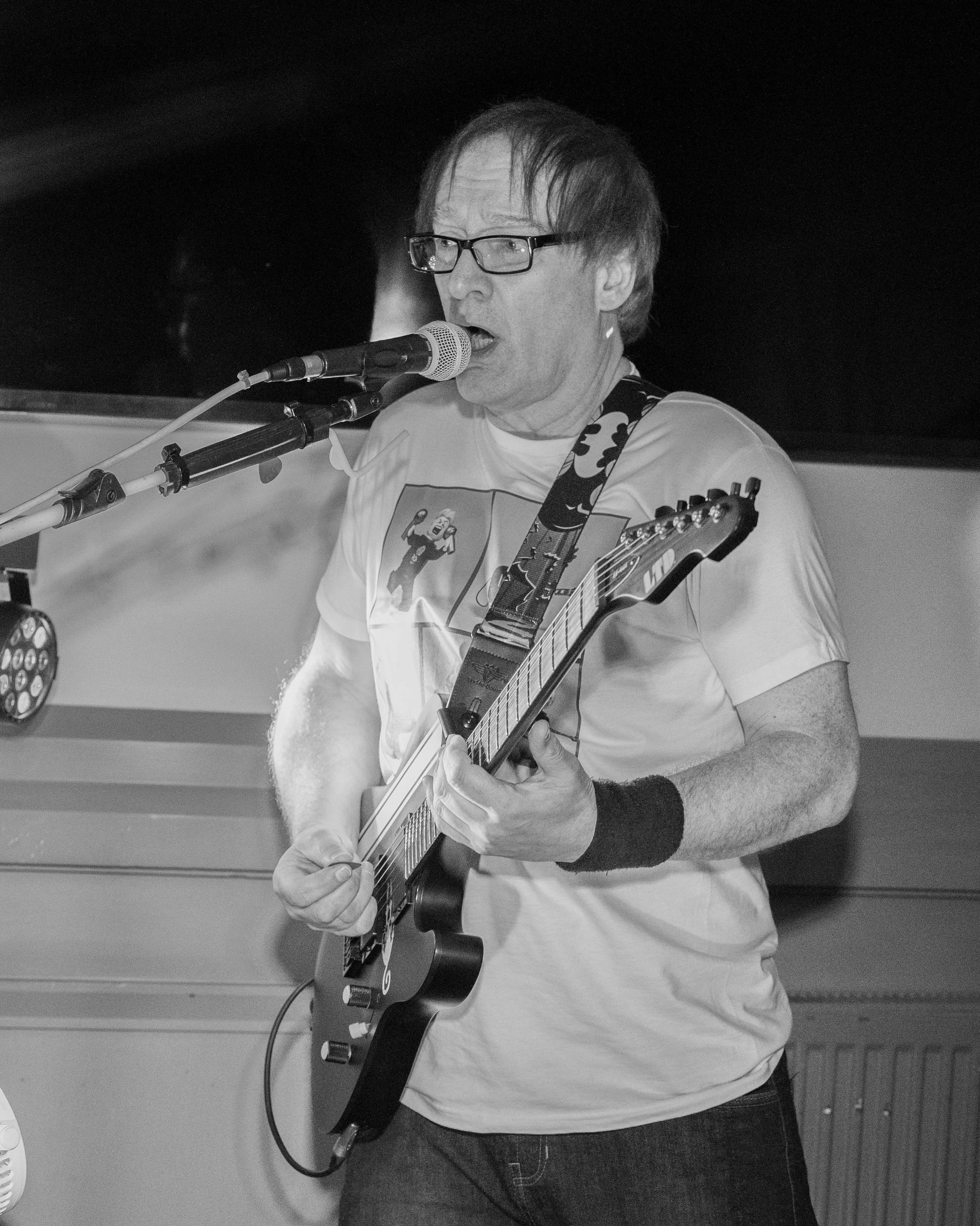 A man wearing glasses and a graphic T-shirt is singing into a microphone while playing an electric guitar.