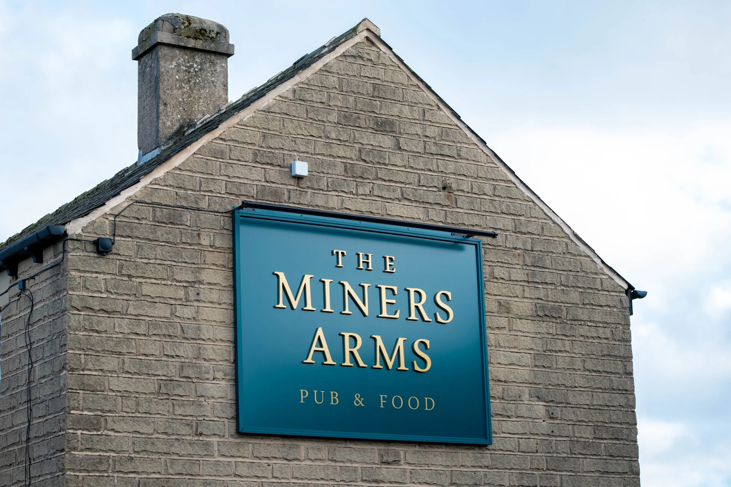 A building with a blue sign that reads "The Miners Arms Pub & Food." The building is made of tan bricks, has a chimney, and a sloped roof.
