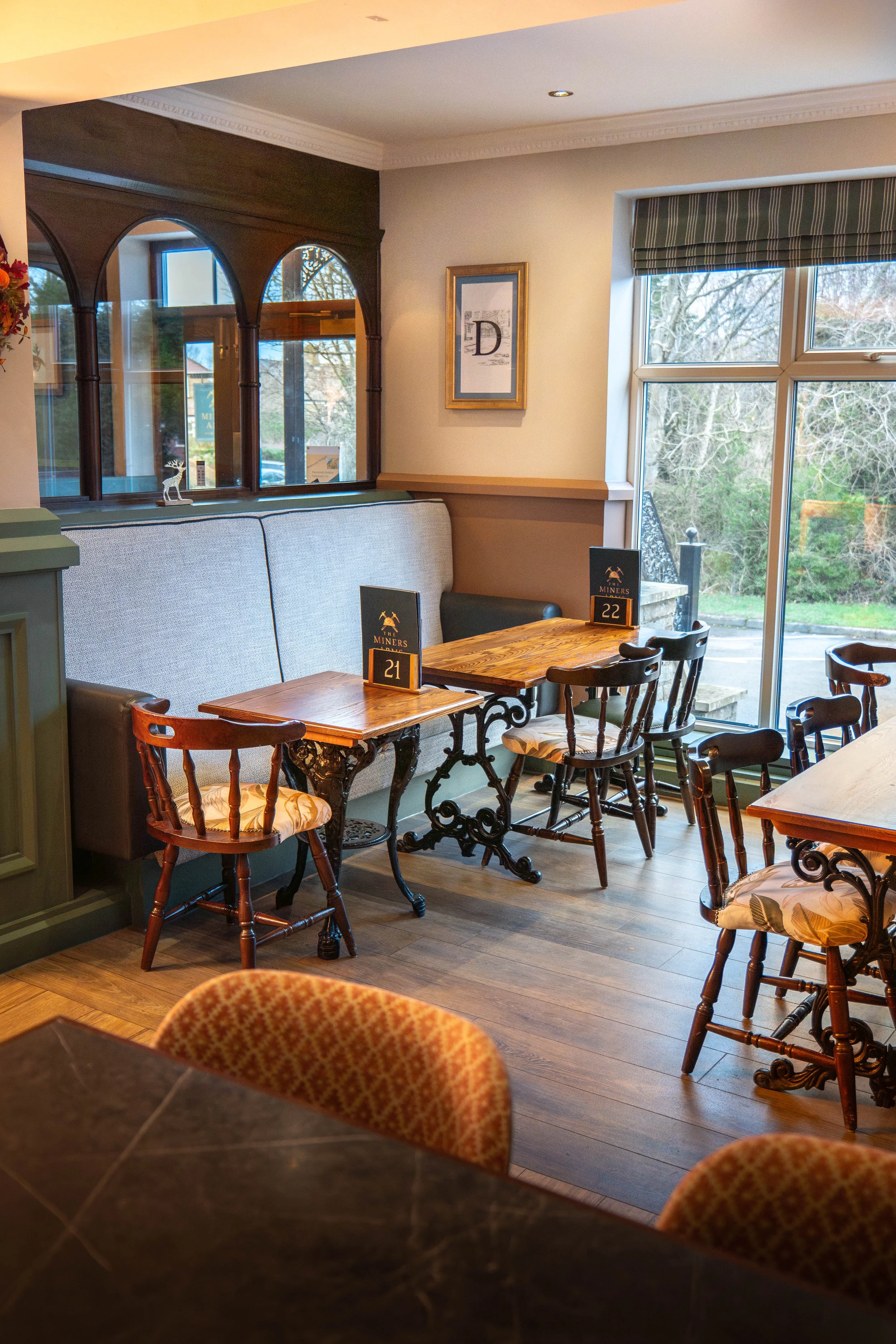 A cozy restaurant corner with wooden tables and chairs, a large window showing trees outside, and table signs numbered 21 and 22.