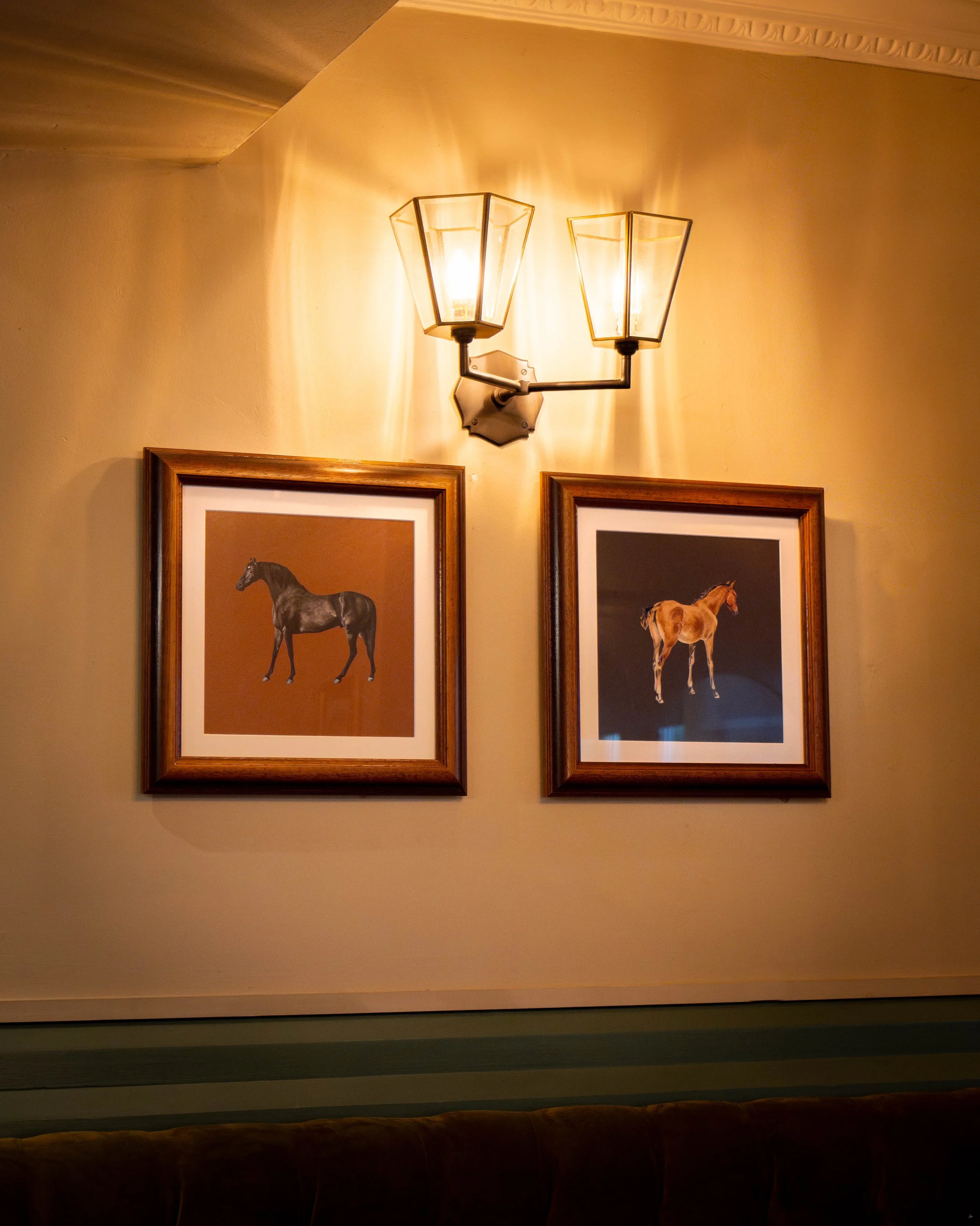 Framed pictures of horses hanging on a beige wall illuminated by a wall-mounted light fixture with two bulbs.