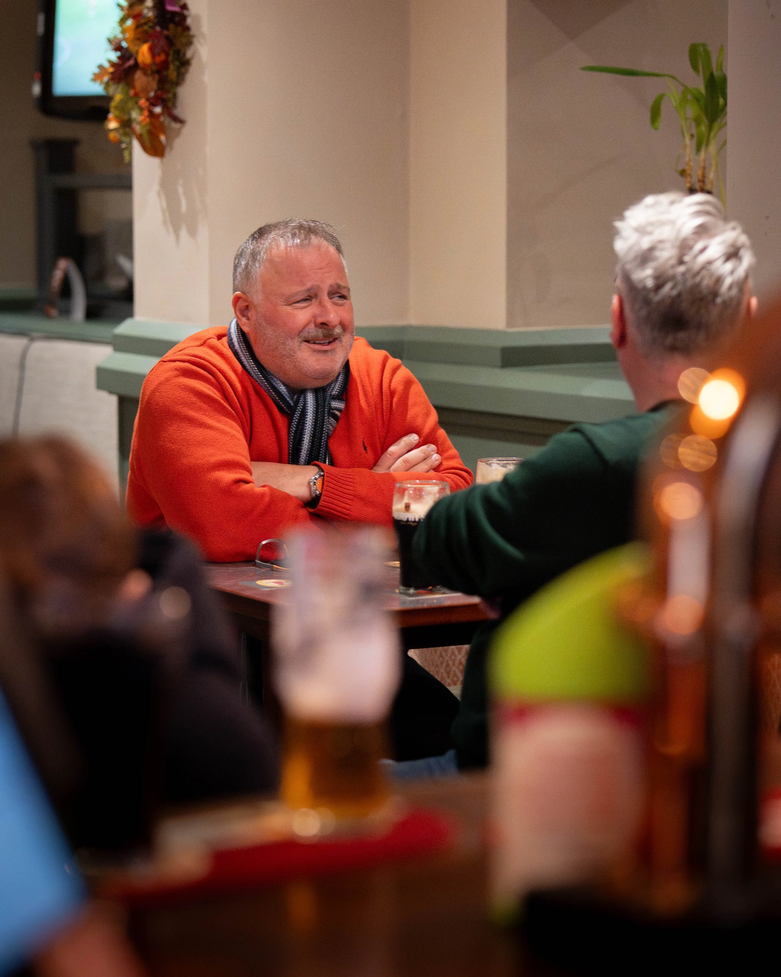 A man wearing an orange sweater and scarf sitting at a restaurant table, crying and looking emotional during a conversation with another person whose back is to the camera.