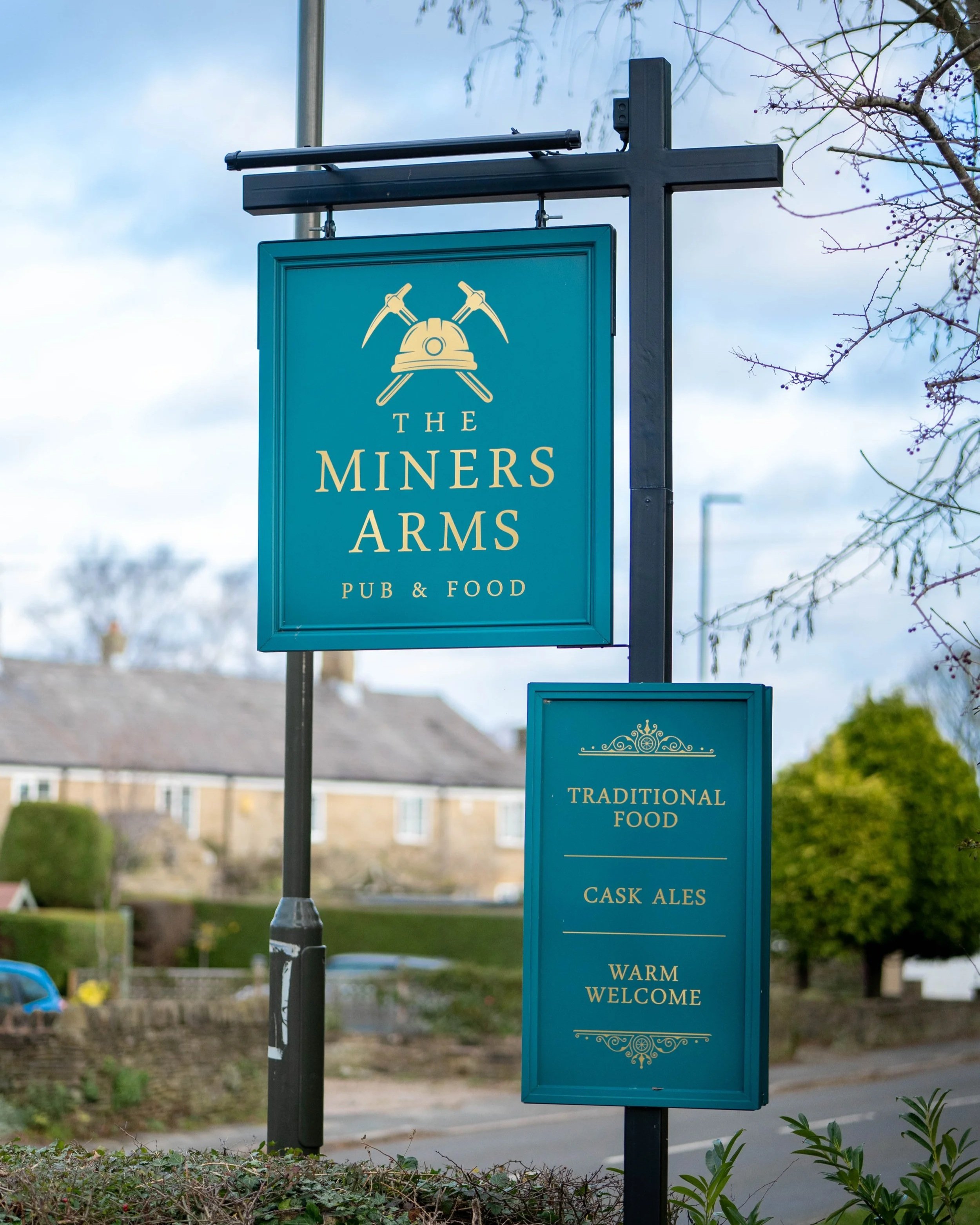 Signage for The Miners Arms pub & food, showing offerings like traditional food, cask ales, warm welcome, with a background of houses, trees, and cloudy sky.