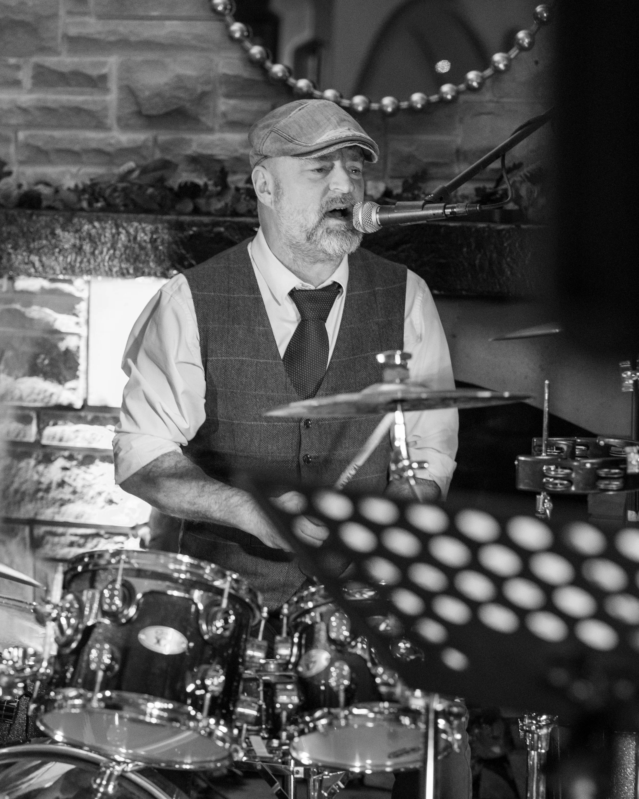 A black and white photo of a man singing into a microphone while playing the drums, wearing a flat cap, vest, shirt, and tie, with a fireplace decorated with garland in the background.
