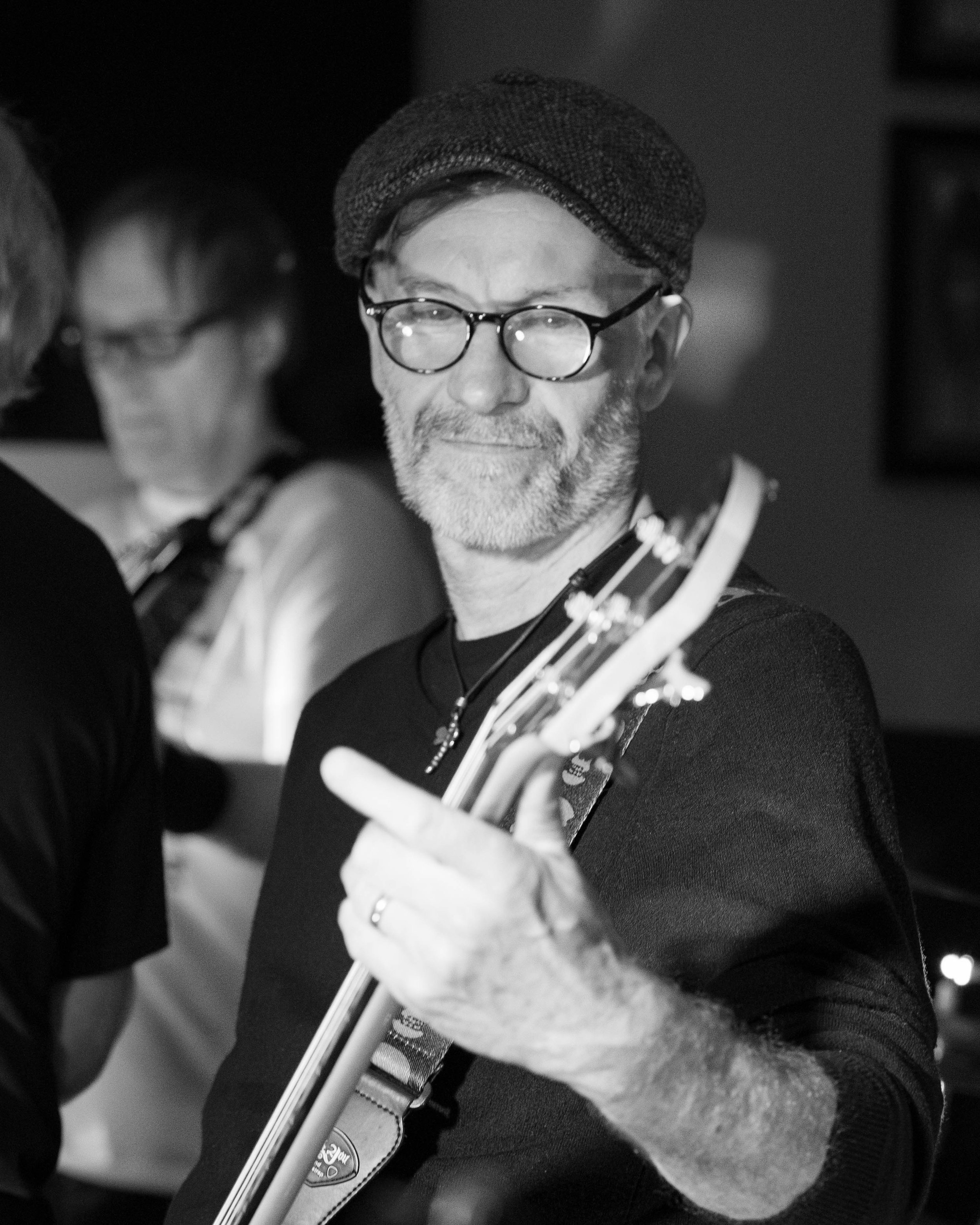 A middle-aged man with glasses, a beard, and a hat smiling while holding a guitar, with other people blurred in the background.
