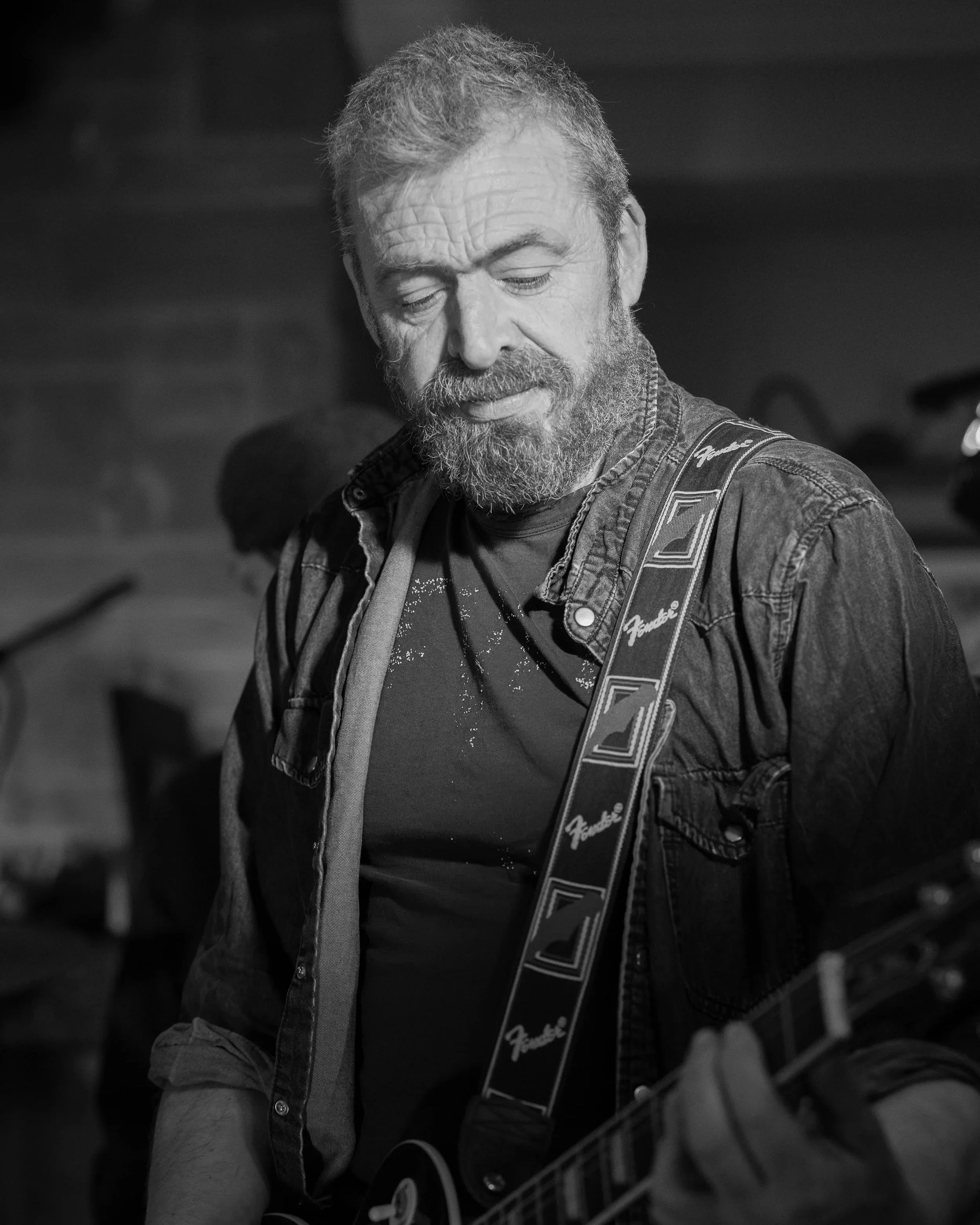 A bearded man with gray hair playing an electric guitar, wearing a jeans jacket and a Fend guitar strap, in a black and white photo.