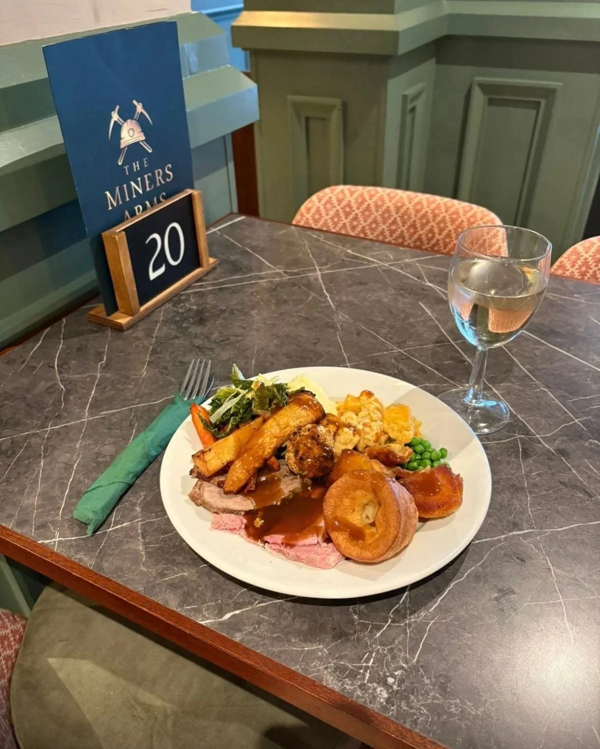 A plate of food with roast beef, vegetables, mashed potatoes, and Yorkshire pudding, on a marble table with a glass of water and a green napkin, at a restaurant called The Miners Arms.
