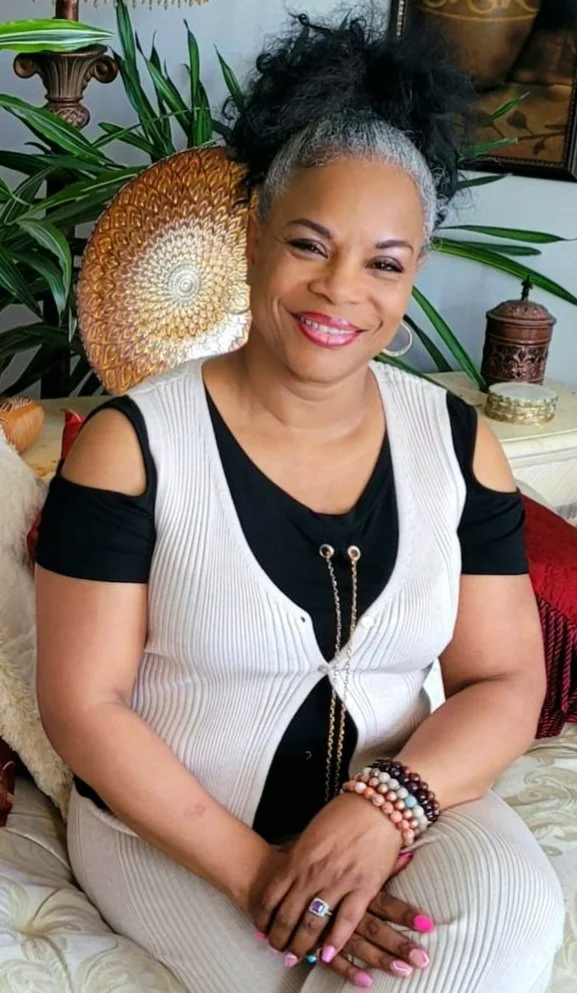 A smiling woman with gray hair styled in a high curly ponytail, wearing a black and white sleeveless top, sitting on a couch with decorative pillows. She has several bracelets and a ring on her left hand. The background includes houseplants, a decorative plate, and a small cabinet.