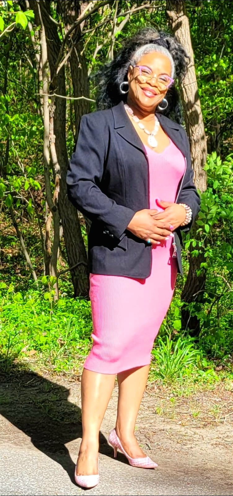 A woman dressed in a pink dress, black blazer, and pink high heels stands outdoors near trees and greenery, smiling and posing for the camera.