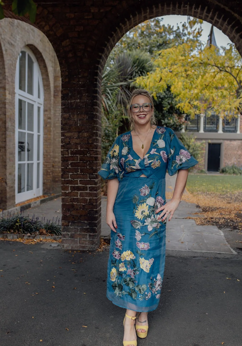 A woman standing outdoors under a brick archway, wearing a blue floral dress and yellow high heels, smiling with glasses and earrings, with autumn trees and a building in the background.