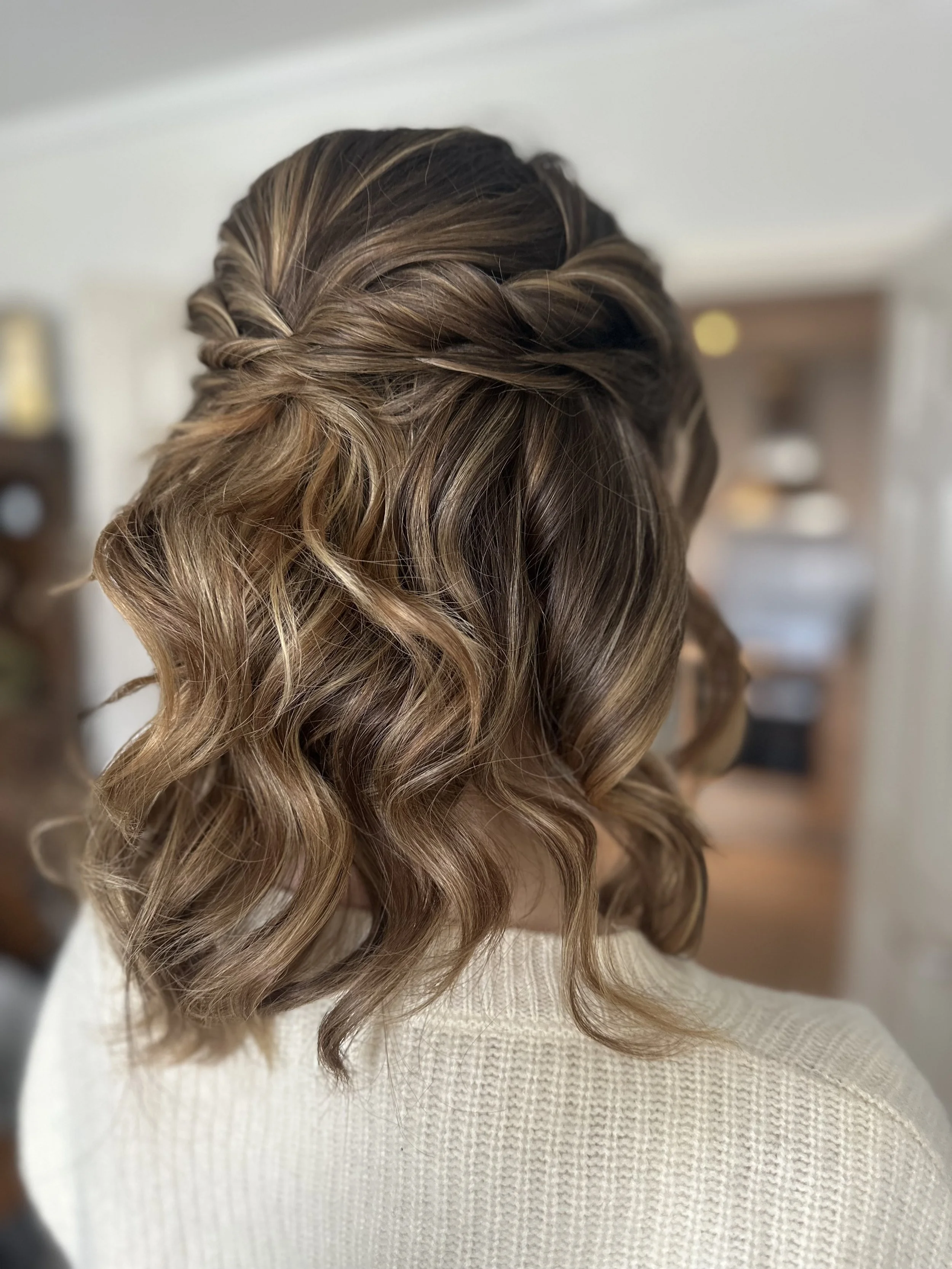 Woman with shoulder-length wavy hair styled with a twist on top, wearing a cream-colored sweater, indoors with blurred shelves in background.