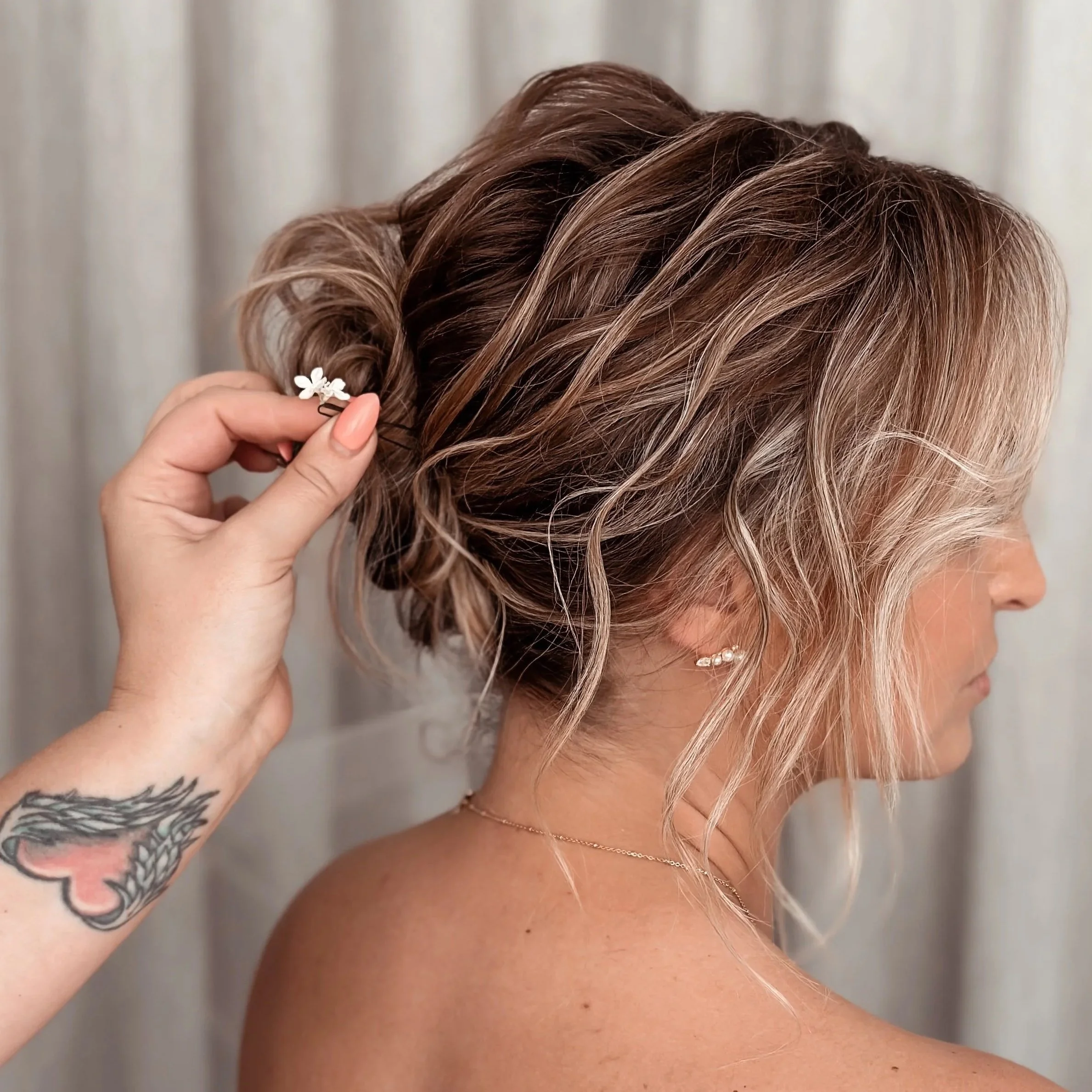 Close-up of a woman with blonde, wavy hair styled in an elegant updo, held in place by a person with a tattoo on their wrist, placing a small floral hair accessory.