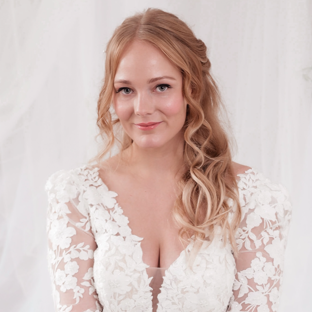 A woman with long, wavy blonde hair wearing a white lace dress, smiling gently against a light background.
