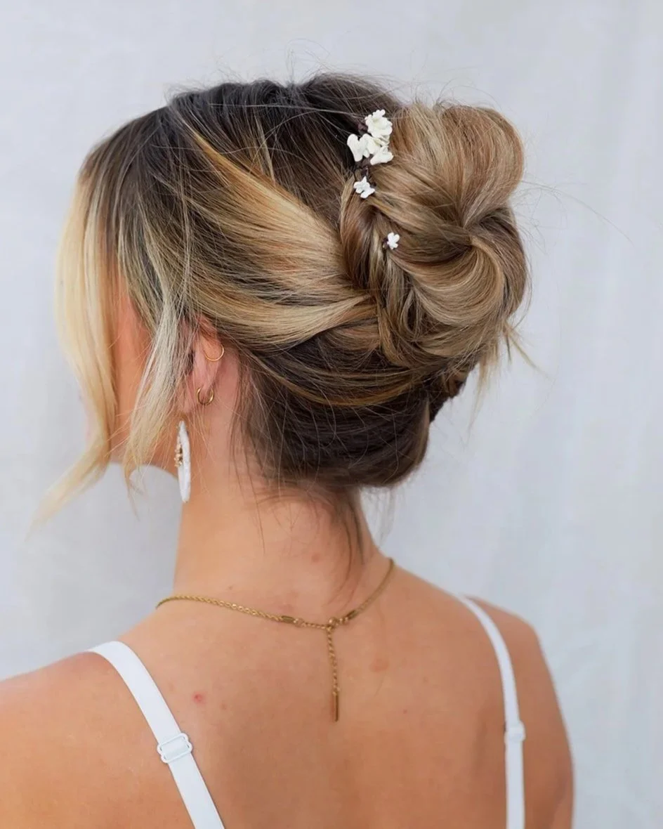 Woman with styled updo hairstyle with small white floral accessories, wearing gold jewelry, and a white spaghetti strap top.