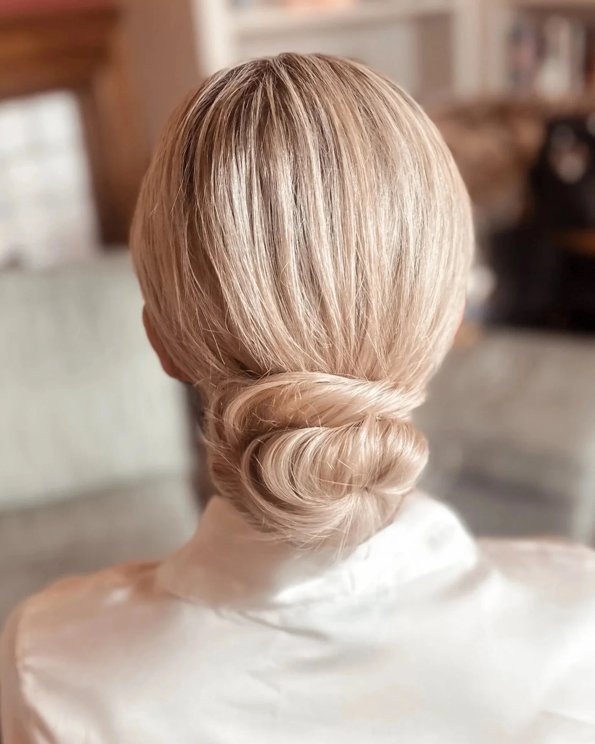 Back of a person's head with styled blonde hair in a low twisted bun, wearing a white satin top, in a cozy indoor setting.