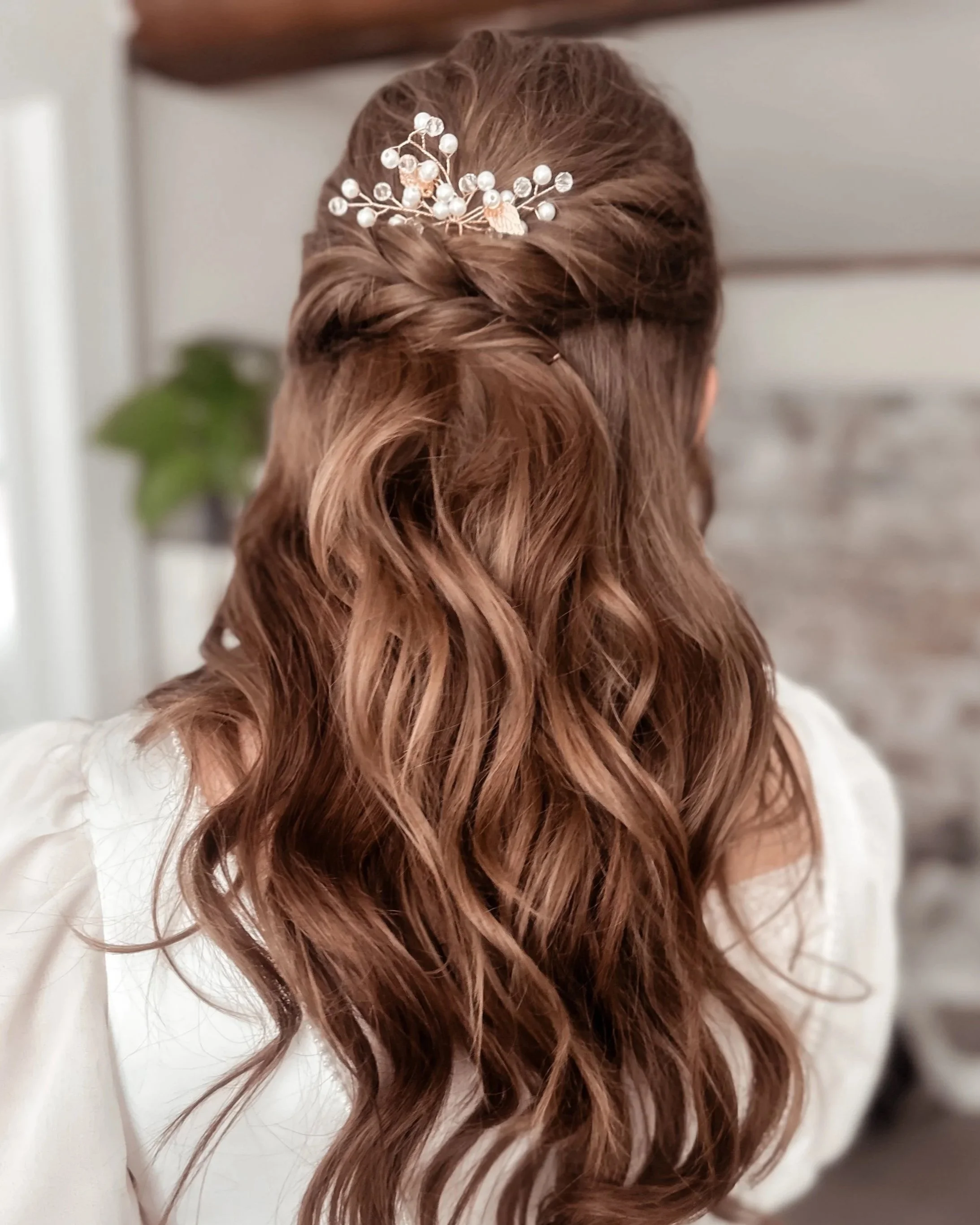 Back of a woman with long, wavy, auburn hair wearing a decorative hair clip with pearls and small flowers, dressed in white.