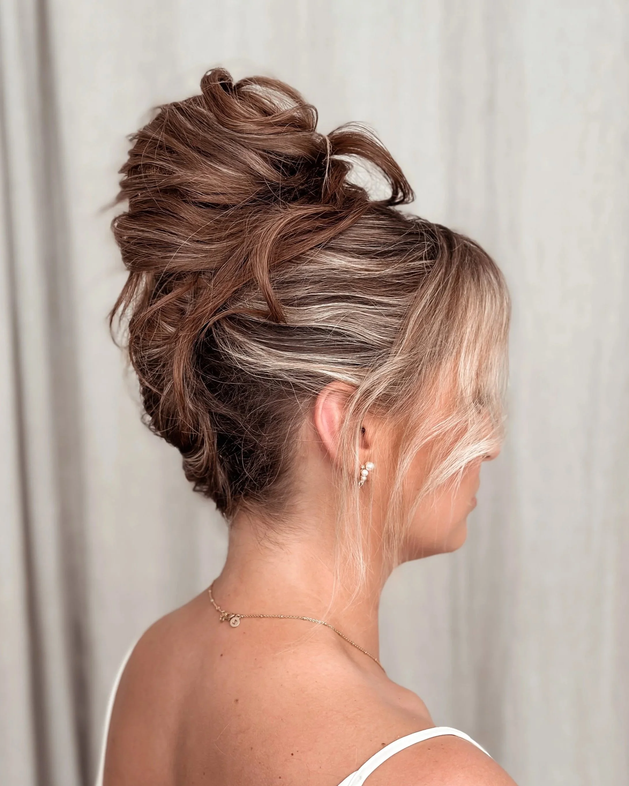 A woman with blonde hair styled in an elegant updo with curled sections, wearing pearl earrings and a delicate necklace, against a light background.