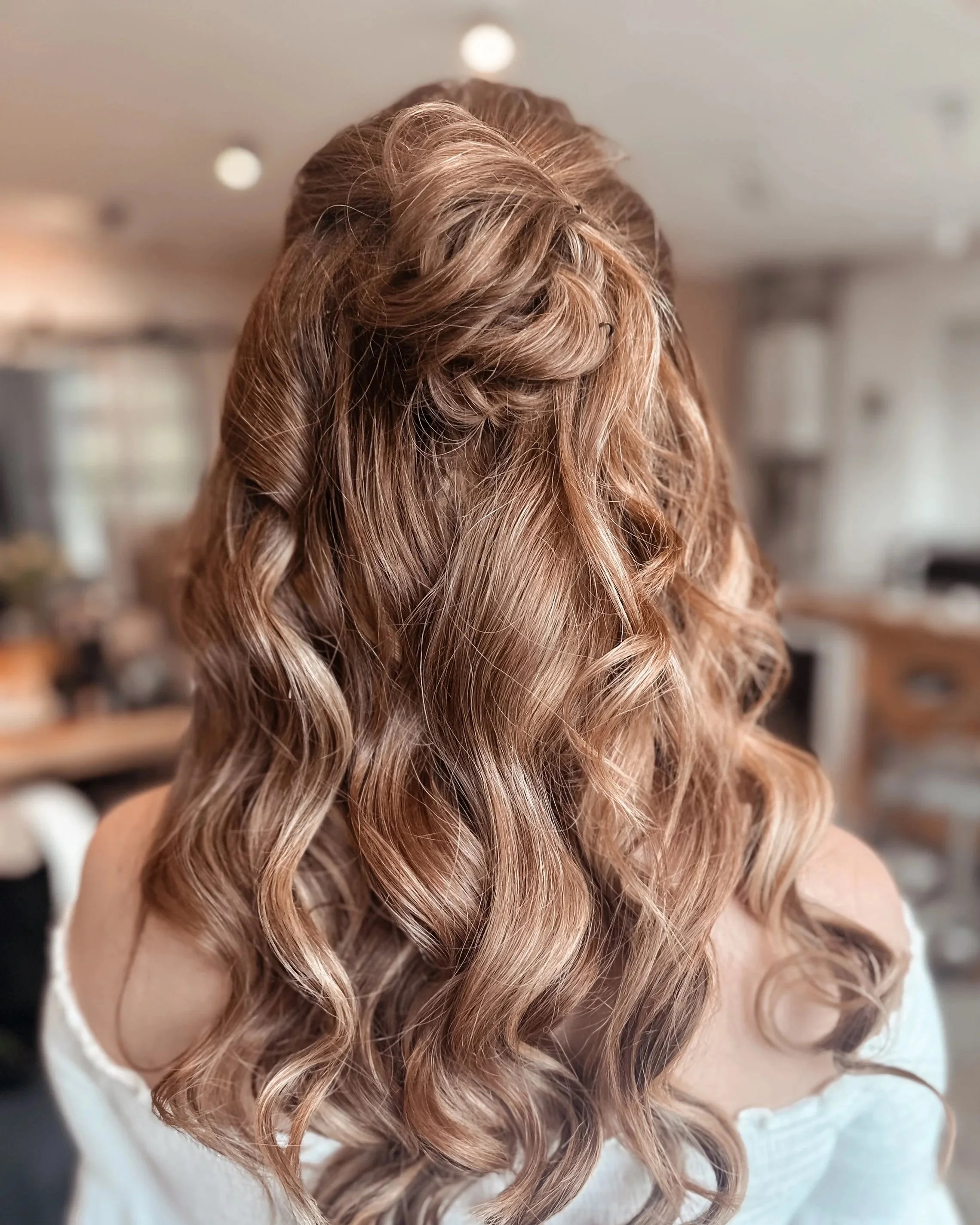 Back view of a woman with long, curly, light brown hair styled with loose waves in a cozy, well-lit room.
