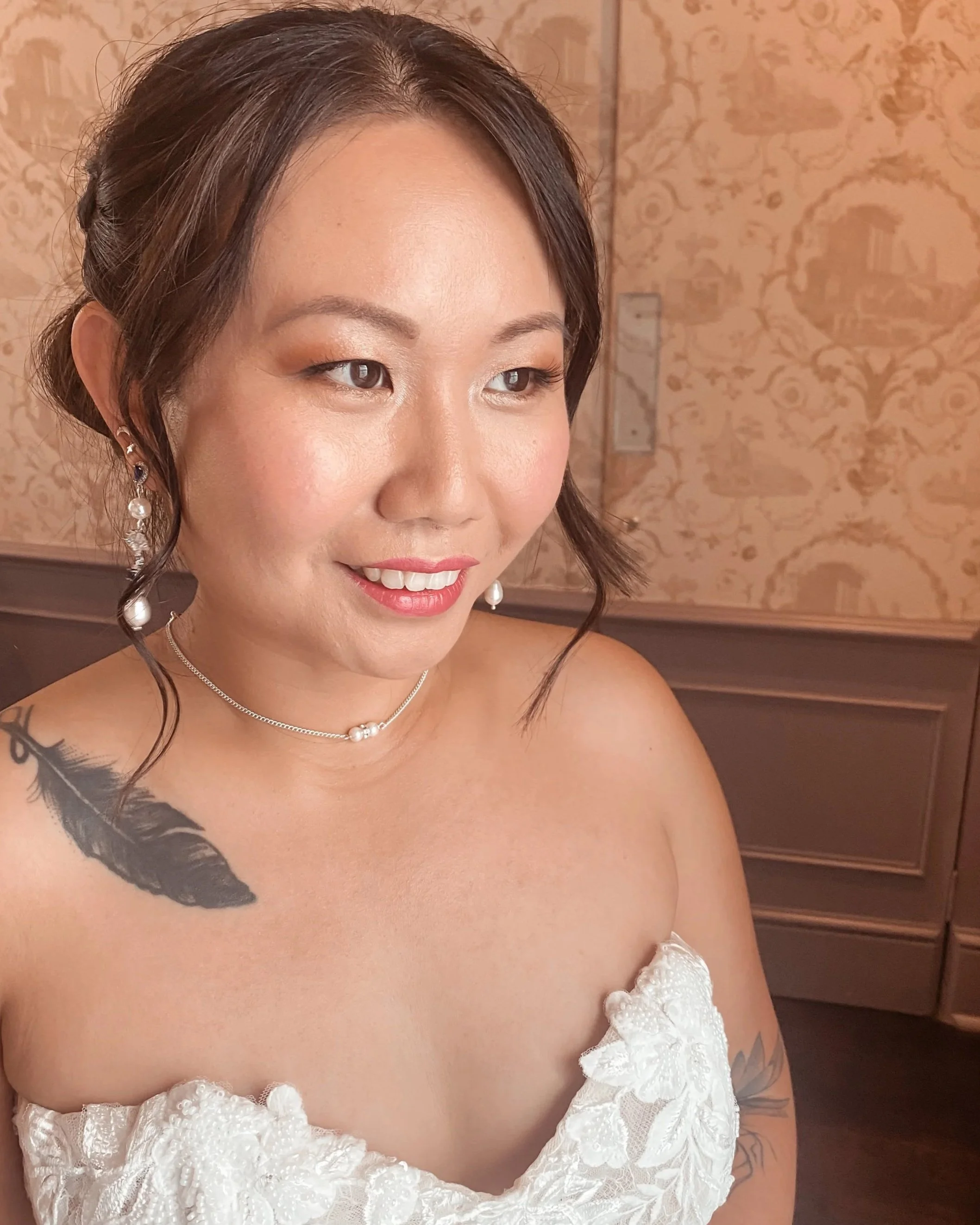 A woman with short dark hair styled in loose waves, wearing pearl and silver jewelry, a white strapless lace dress, and showing tattoos of a feather and a flower on her shoulder.