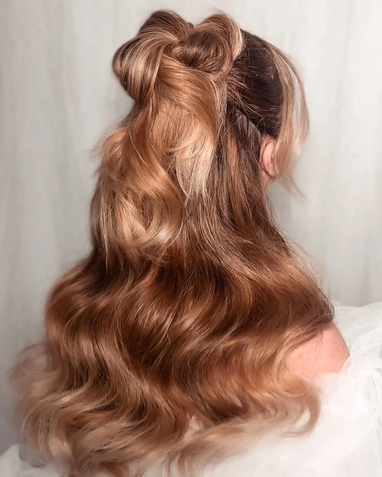 Woman's head turned away, showcasing long, wavy, caramel-colored hair styled with an intricate updo near the crown and loose curls flowing down.