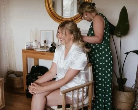 A bride sits on a wooden chair smiling as another hair and   makeup artist styles her hair in a room with a large mirror, a dresser, and a tall plant.