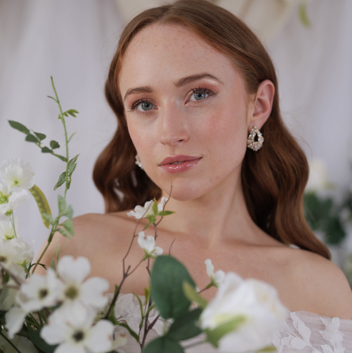 Close-up portrait of a young woman with red hair and blue eyes, wearing pearl earrings, surrounded by white flowers and green leaves.