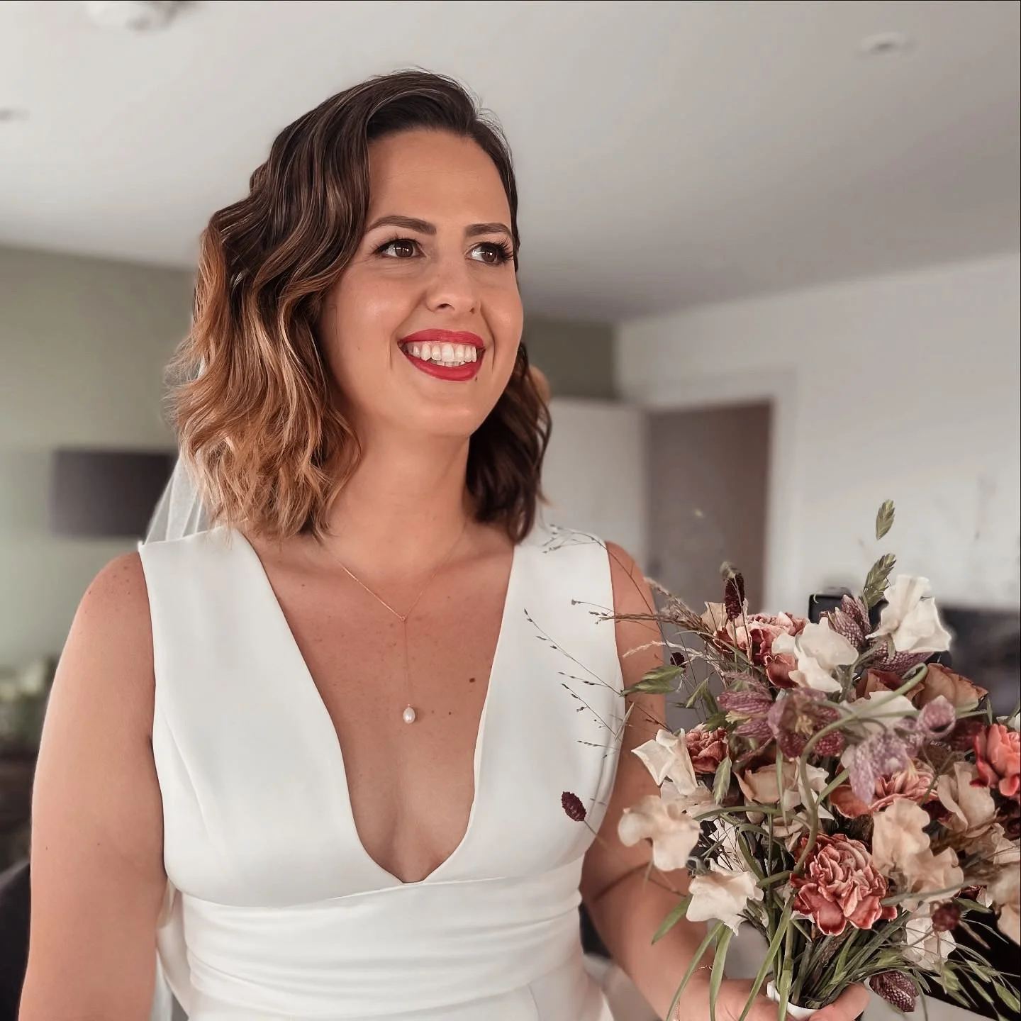A smiling woman with shoulder-length wavy brown hair in a white dress holding a bouquet of pink and cream flowers.