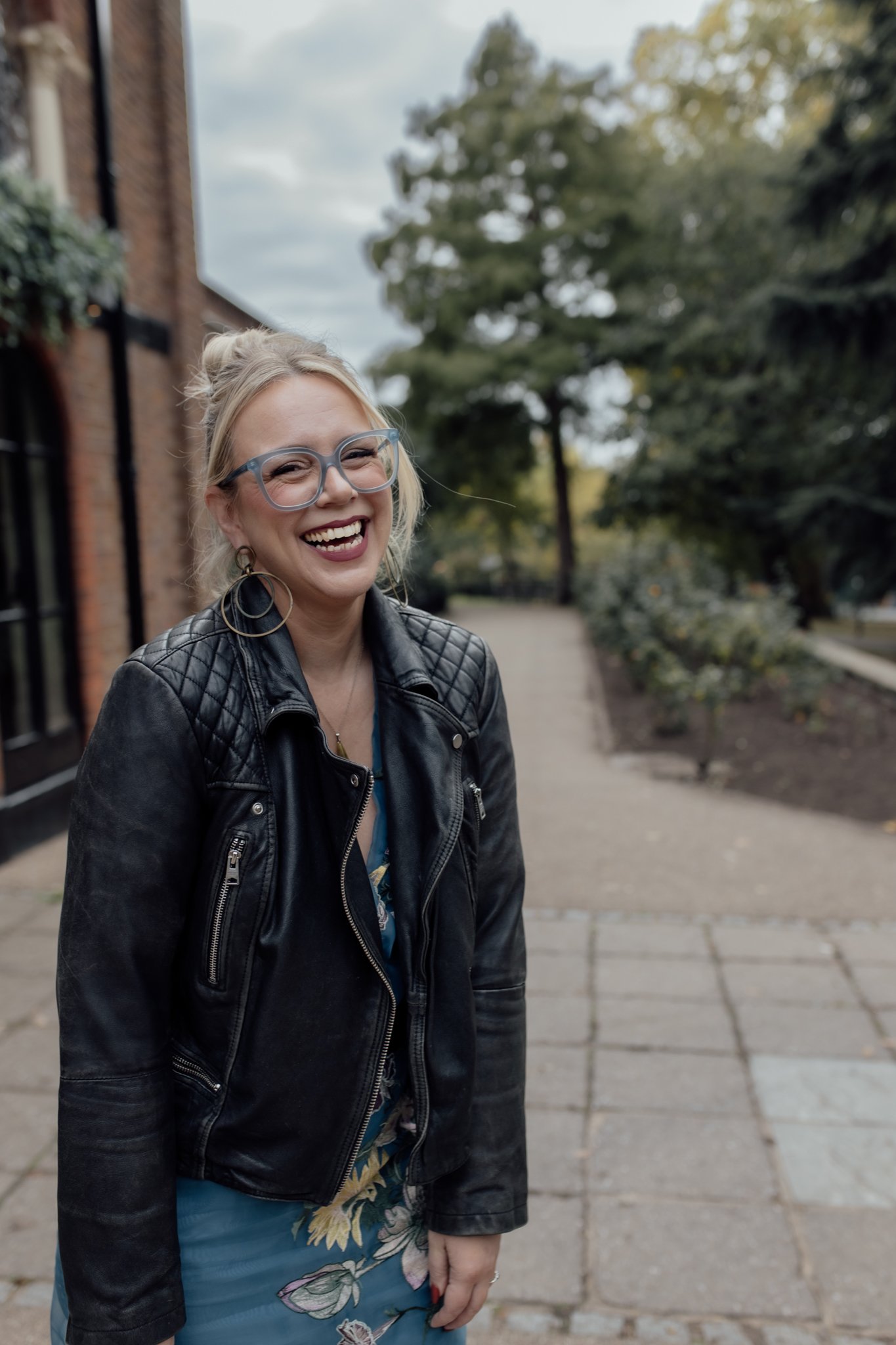 A woman with blonde hair, glasses, and hoop earrings, wearing a black leather jacket, smiling outdoors on a sidewalk with trees in the background.