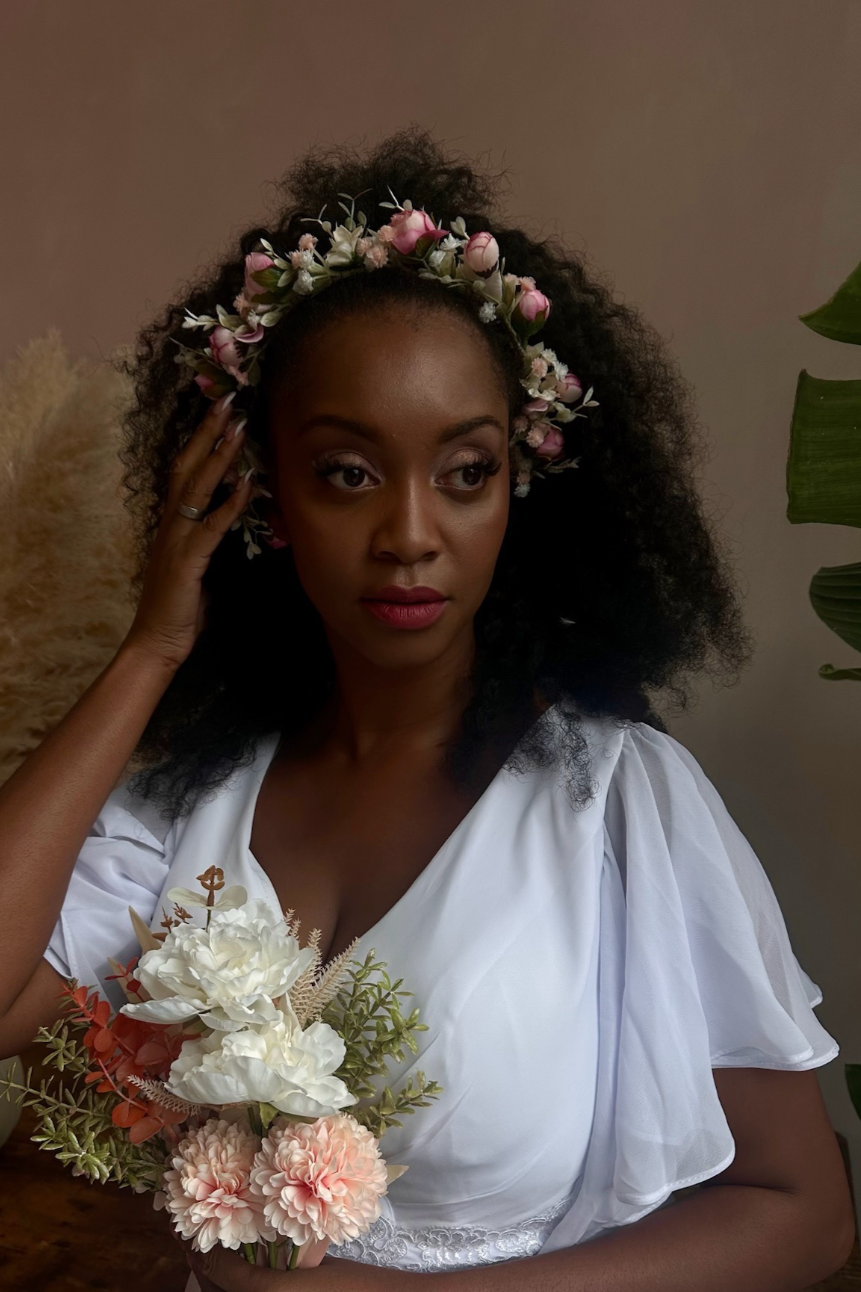 A black woman with dark curly hair wearing a floral crown, a white dress with puffed sleeves, holding a bouquet of white and pink flowers, and standing indoors next to green plants.