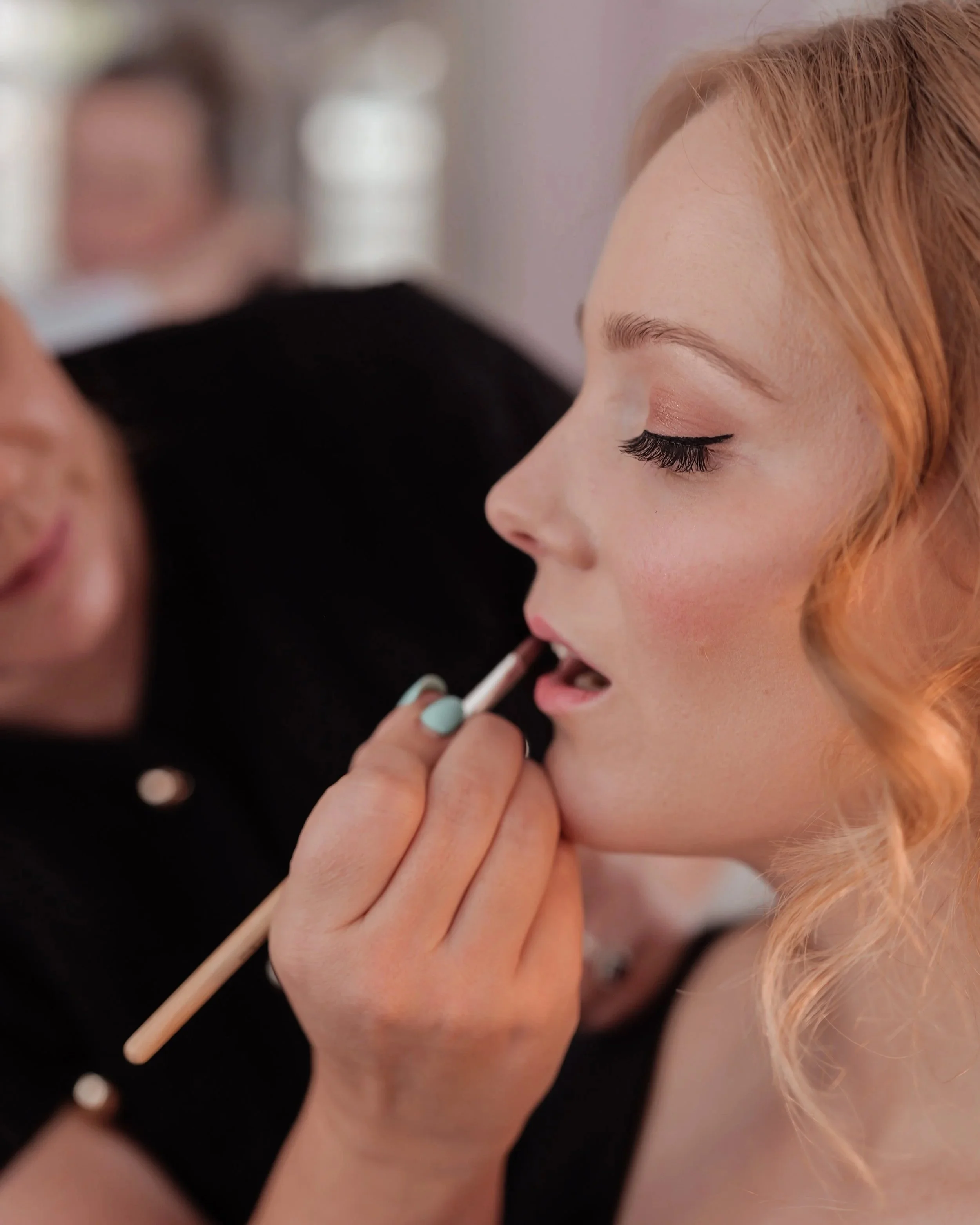 A woman with light hair getting her lips painted by a makeup artist.