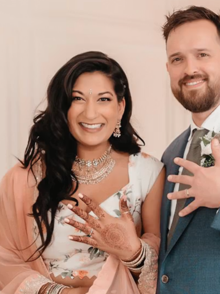 A smiling woman wearing soft glam makeup traditional Indian jewellery and attire showing her wedding hennaed hand, standing beside a man in a suit, both showing off their rings.