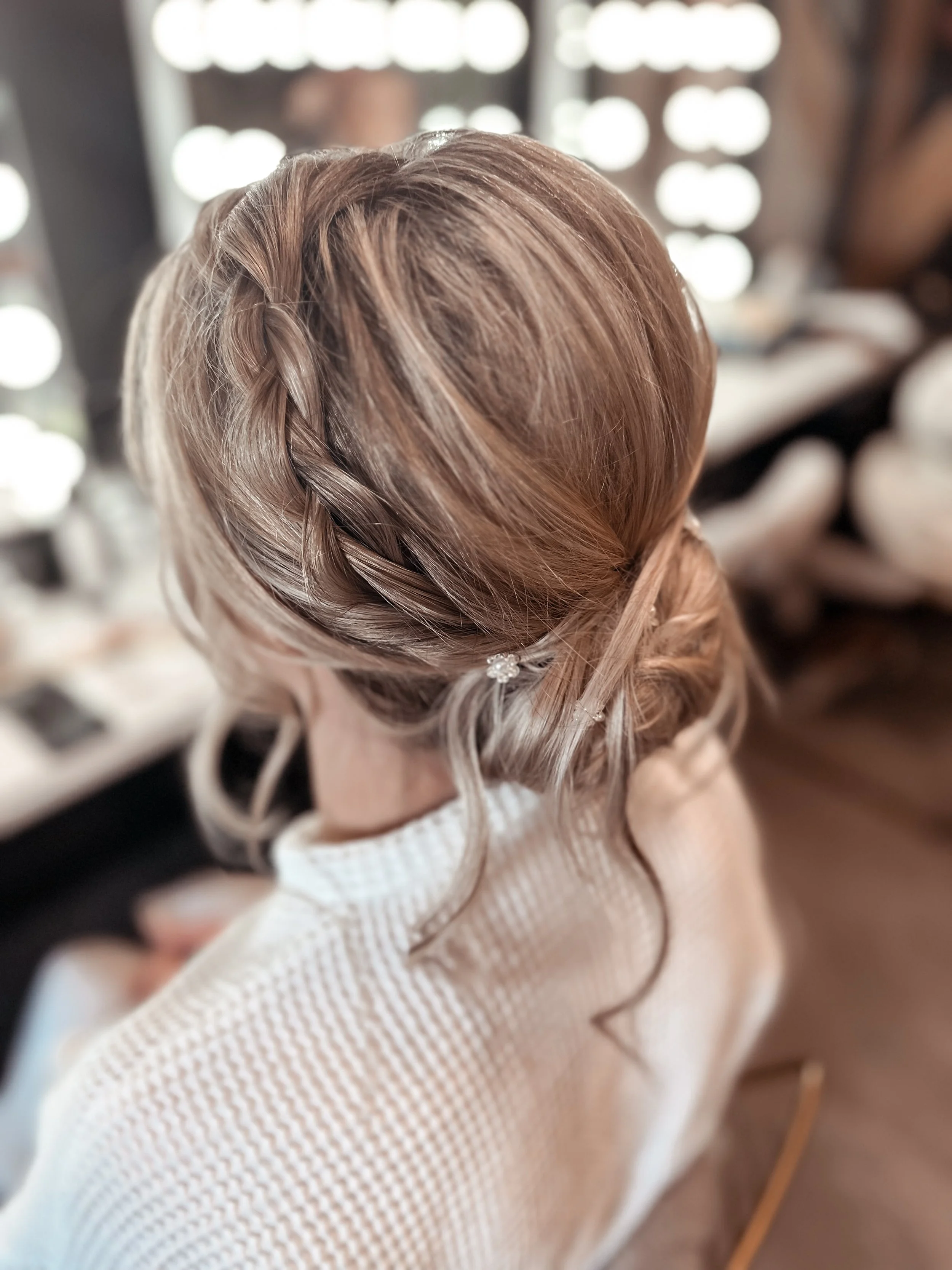 A woman with styled blonde hair, featuring a braid and loose curls, sitting at a likely beauty salon, with makeup stations and people blurred in the background.