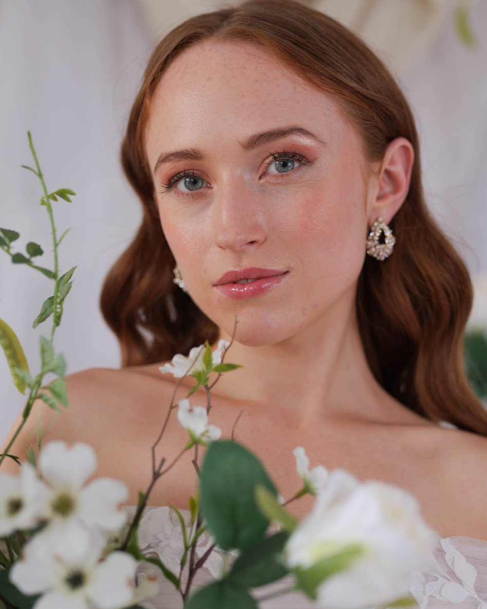 A young woman with natural makeup red hair and blue eyes wearing pearl earrings, surrounded by white flowers and green leaves.