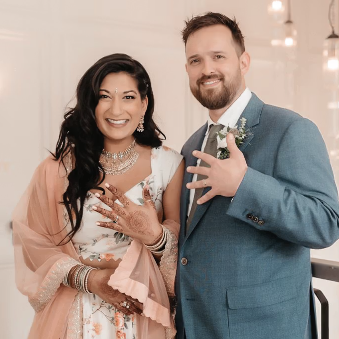 A joyful bride and groom, dressed in wedding attire, showing off rings on their fingers in a well-lit indoor setting.