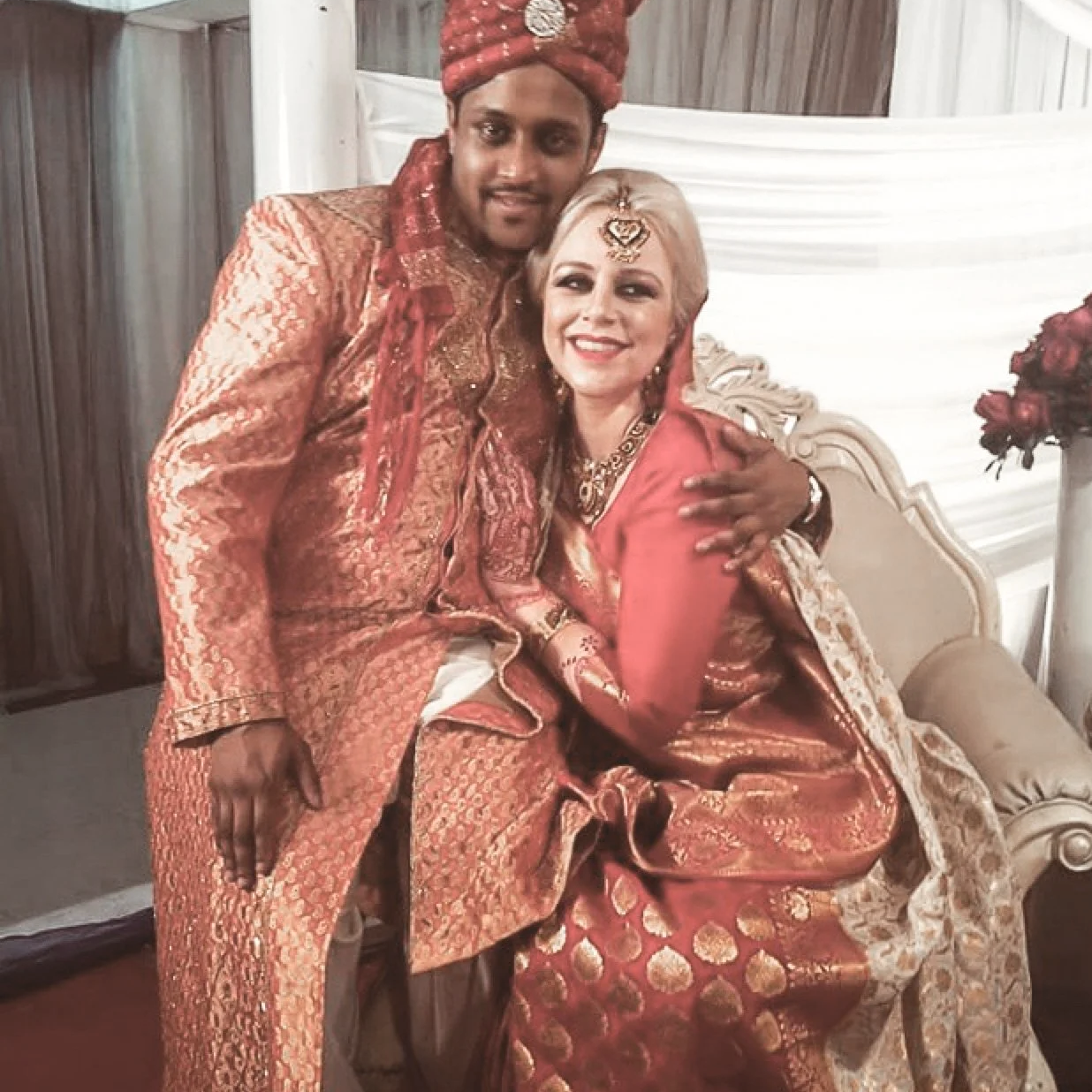 A man and woman dressed in traditional Indian wedding attire, sitting and standing together, smiling at a wedding celebration.