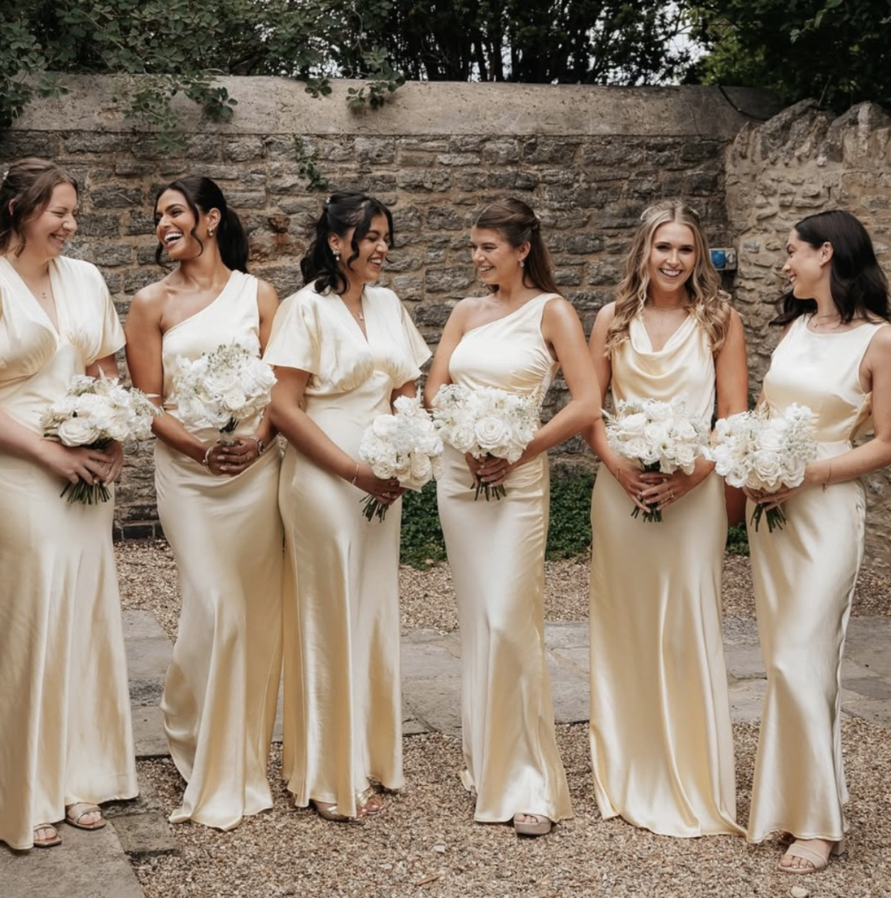 A group of six women dressed in matching cream-colored satin dresses holding white flower bouquets, standing outdoors against a brick wall, smiling and laughing.